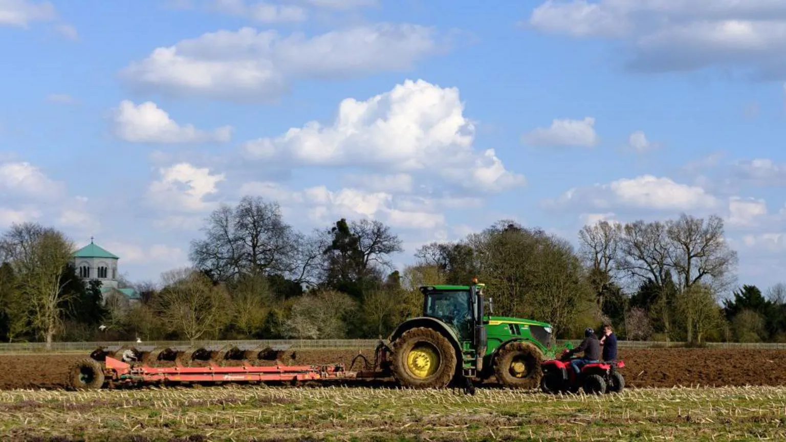  A green tractor in a field of grass and brown earth, with bare but green trees in the background and a blue sky with white clouds. Next to the tractor are two people on a red quad bike