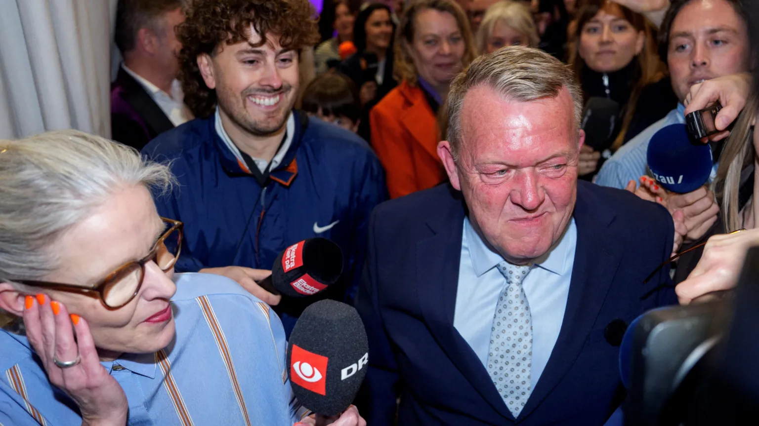  Party leader Lars Loekke Rasmussen makes his way through a crowd of journalists holding microphones and cameras. He is wearing a suit and smiling. 
