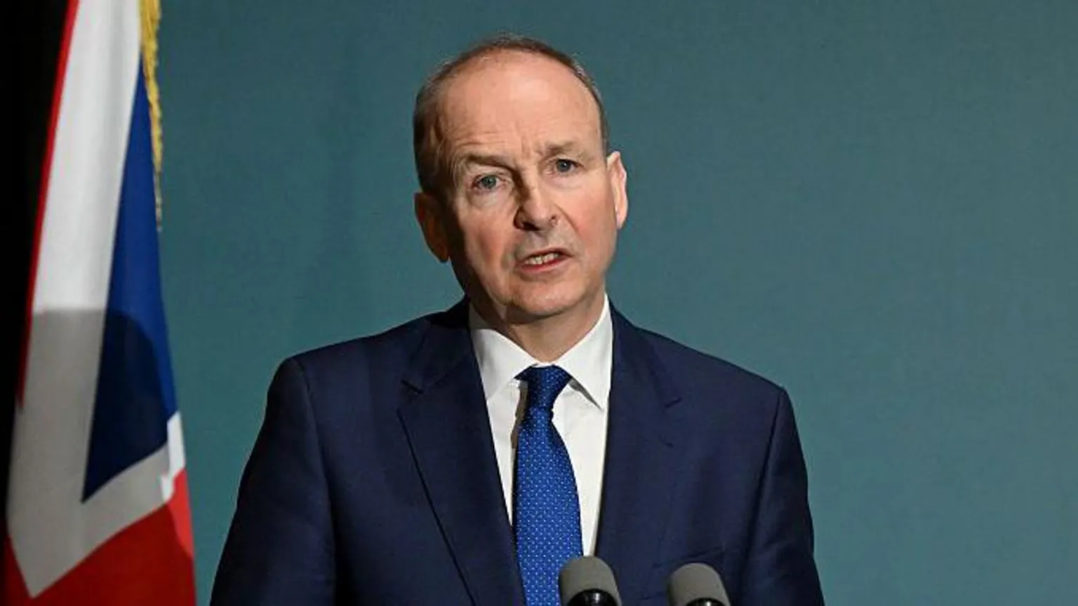  Micheál Martin speaking into a microphone. He is wearing a navy suit, white shirt and a blue tie.