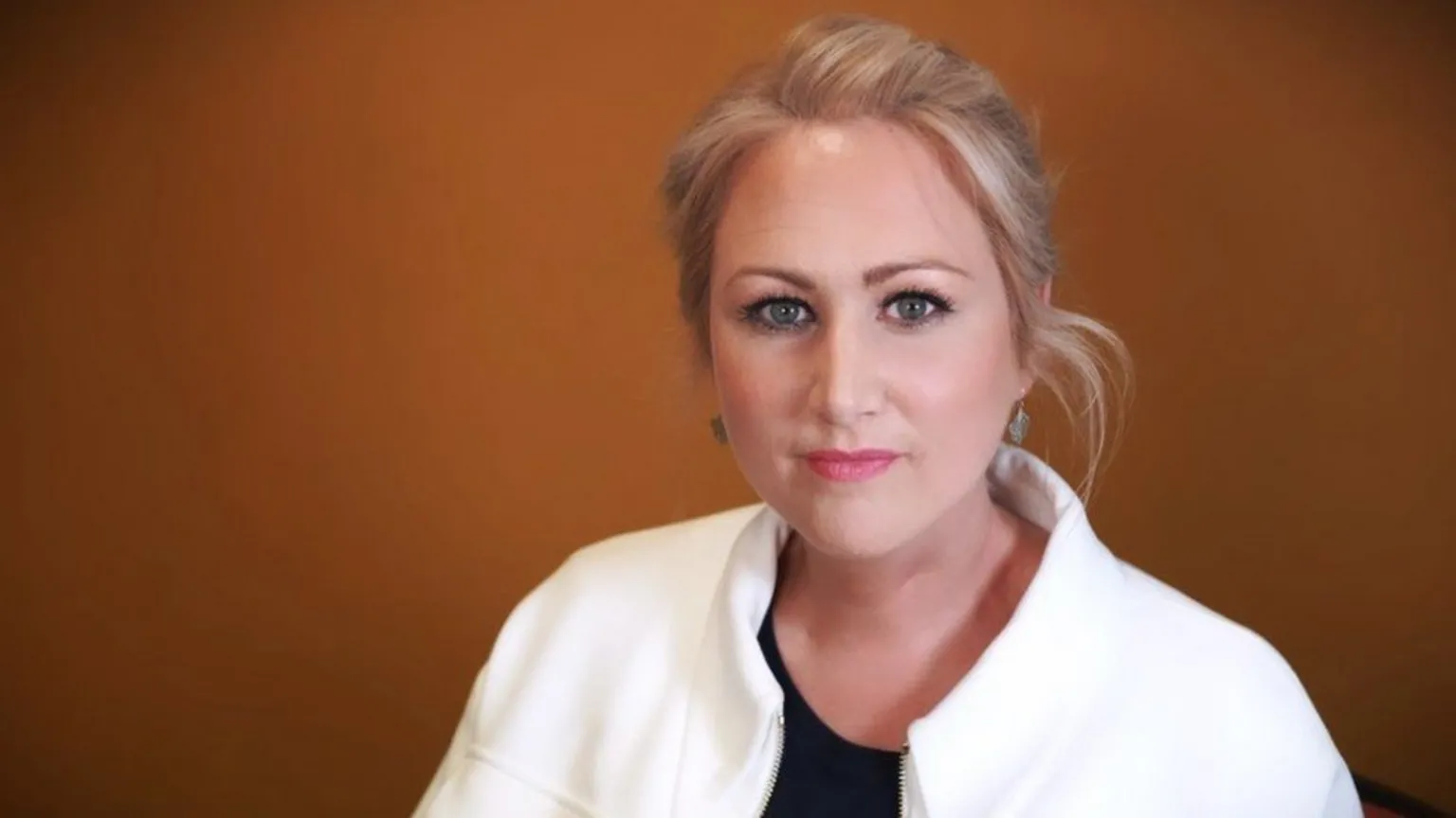 S4C Llinos Griffin-Williams standing in front of an orange background. She is looking at the camera. She has her blonde hair tied back and is wearing a white jacket and a black round neck top.