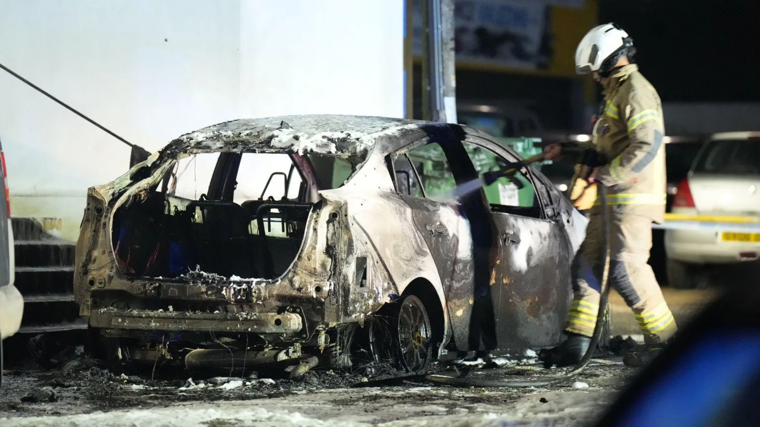Eddie Mitchell A firefighter spraying a hose into the right rear window of a burned car at night. The photo is taken from behind and to the right of the vehicle