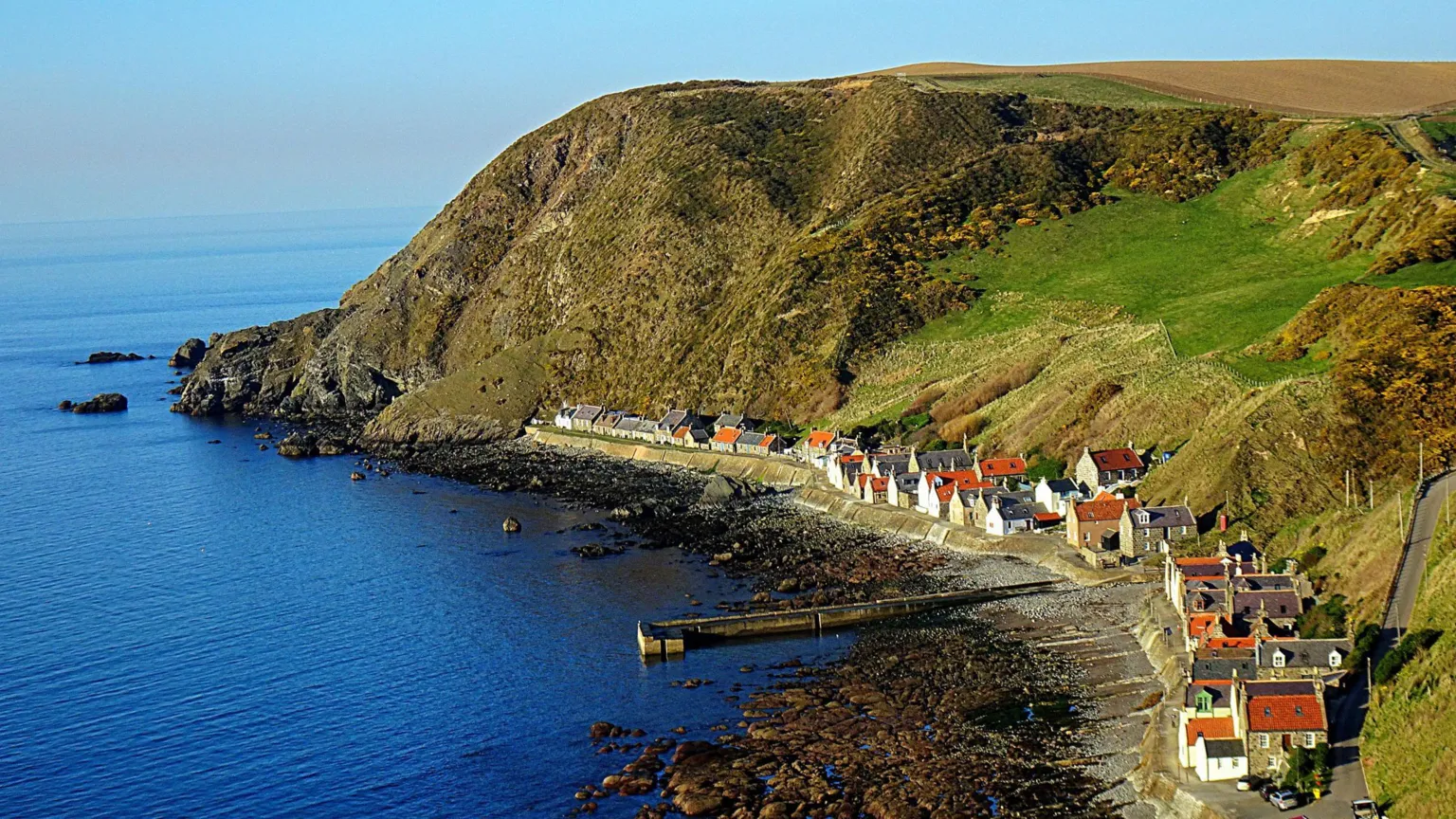 Saf37y/BBC Weather Watchers A view down to the coastal village of Crovie. The row of white-walled houses with orange roofs are at the bottom of a steep, grassy cliff. The tide is out revealing a rocky shoreline.