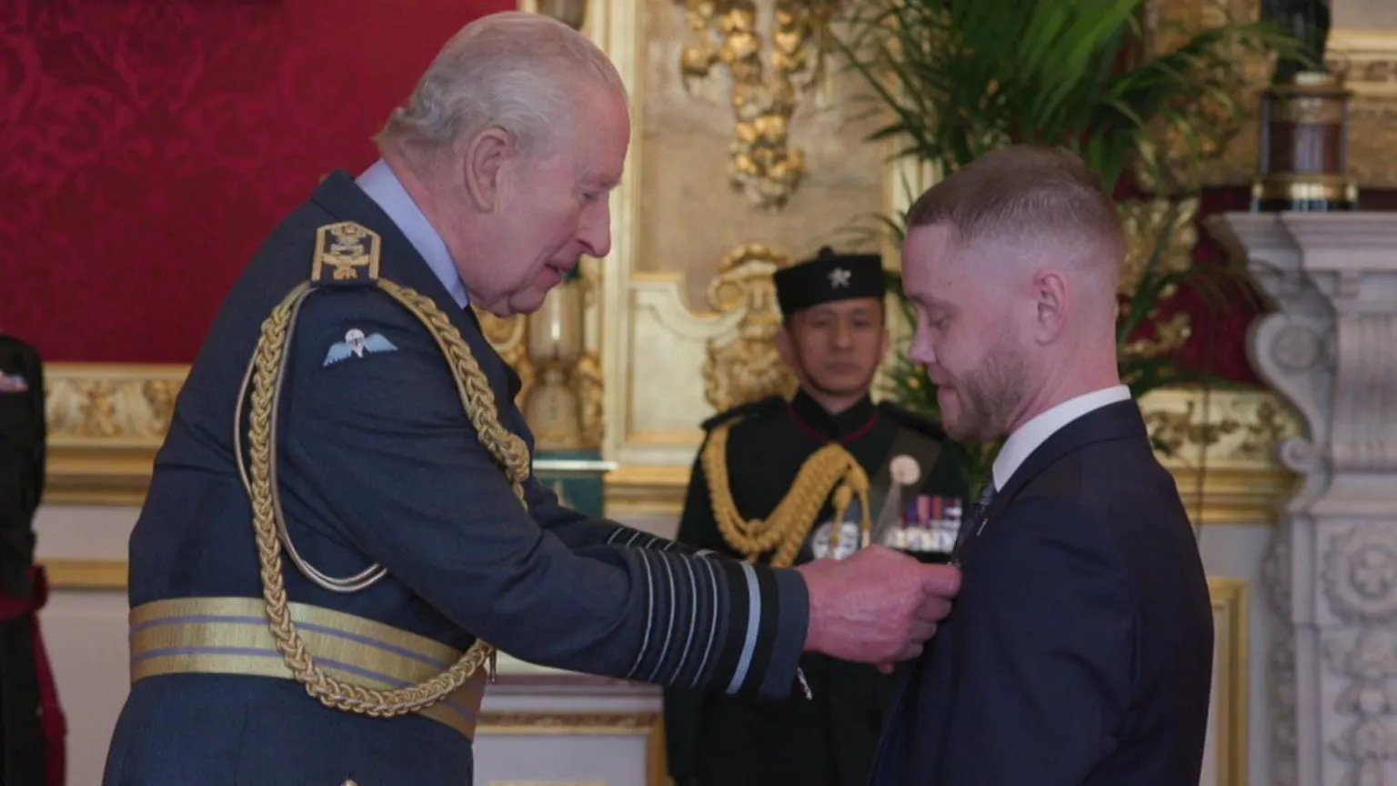 BRITISH CEREMONIAL ARTS (BCA)/Nathan Newby King Charles in RAF uniform smiles at Nathan Newby, a man in a dark blue suit, as he pins a medal on him. In the background is another man in military uniform who stands in a grand-looking room with gold decorations and a carved marble fireplace.