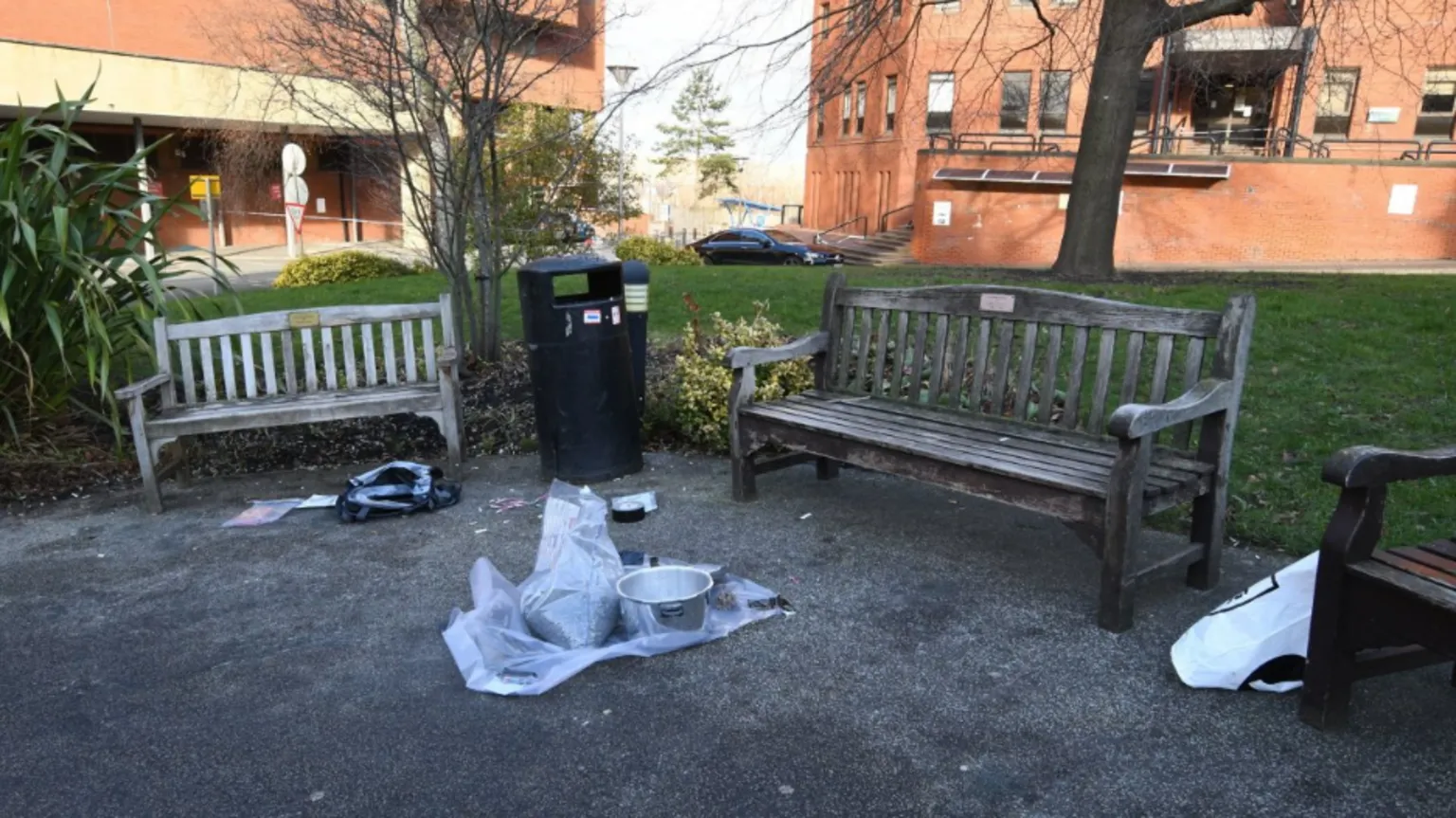 PA A seating area outside the hospital. Three wooden benches are on gravel. A presser cooker with no lid sits on a sheet of material. Bags are scattered around. 