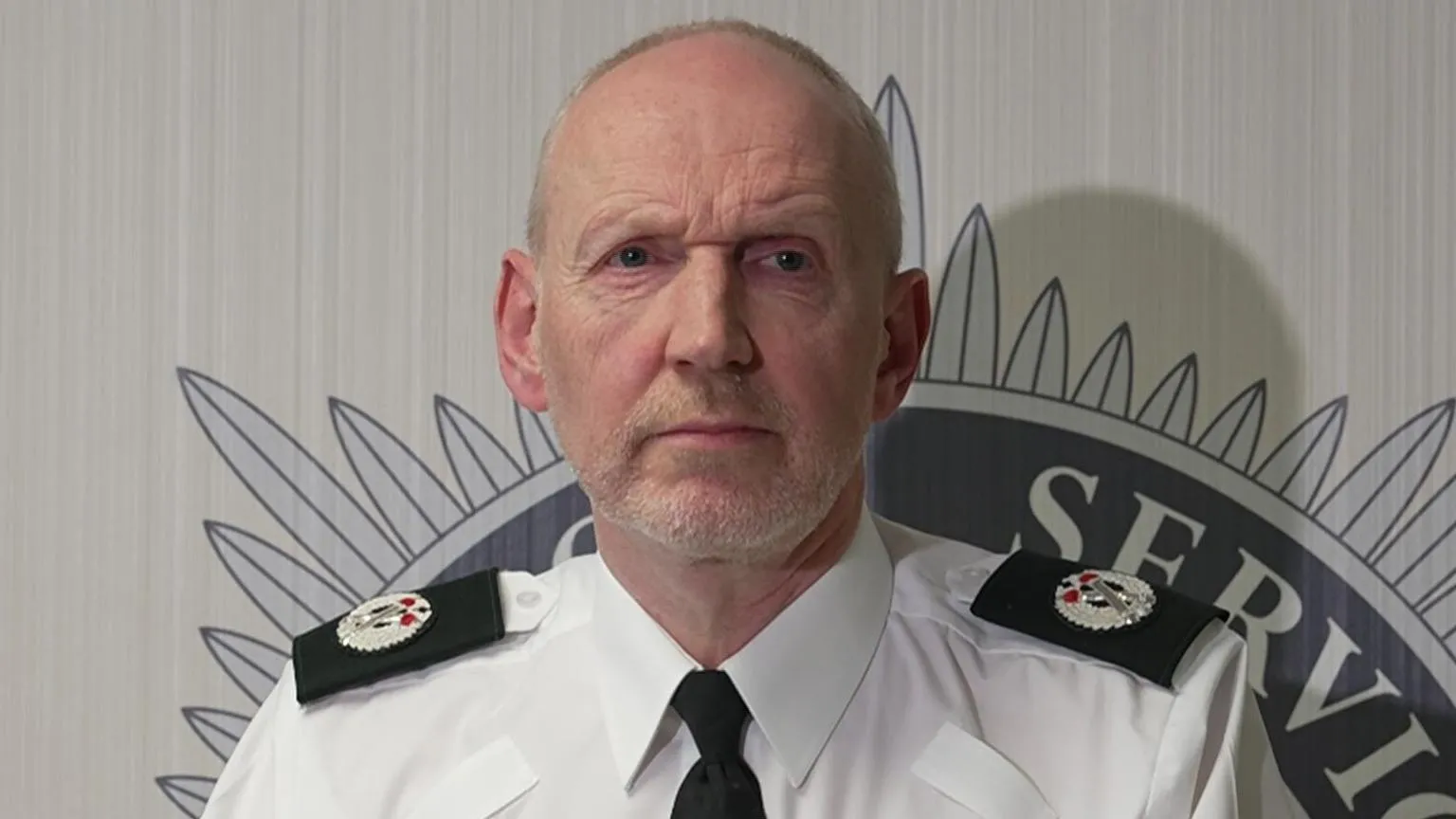 A still of Davy Beck at a press conference. He is an older, bald man with a white beard. He is wearing a police uniform and has a neutral expression on his face.