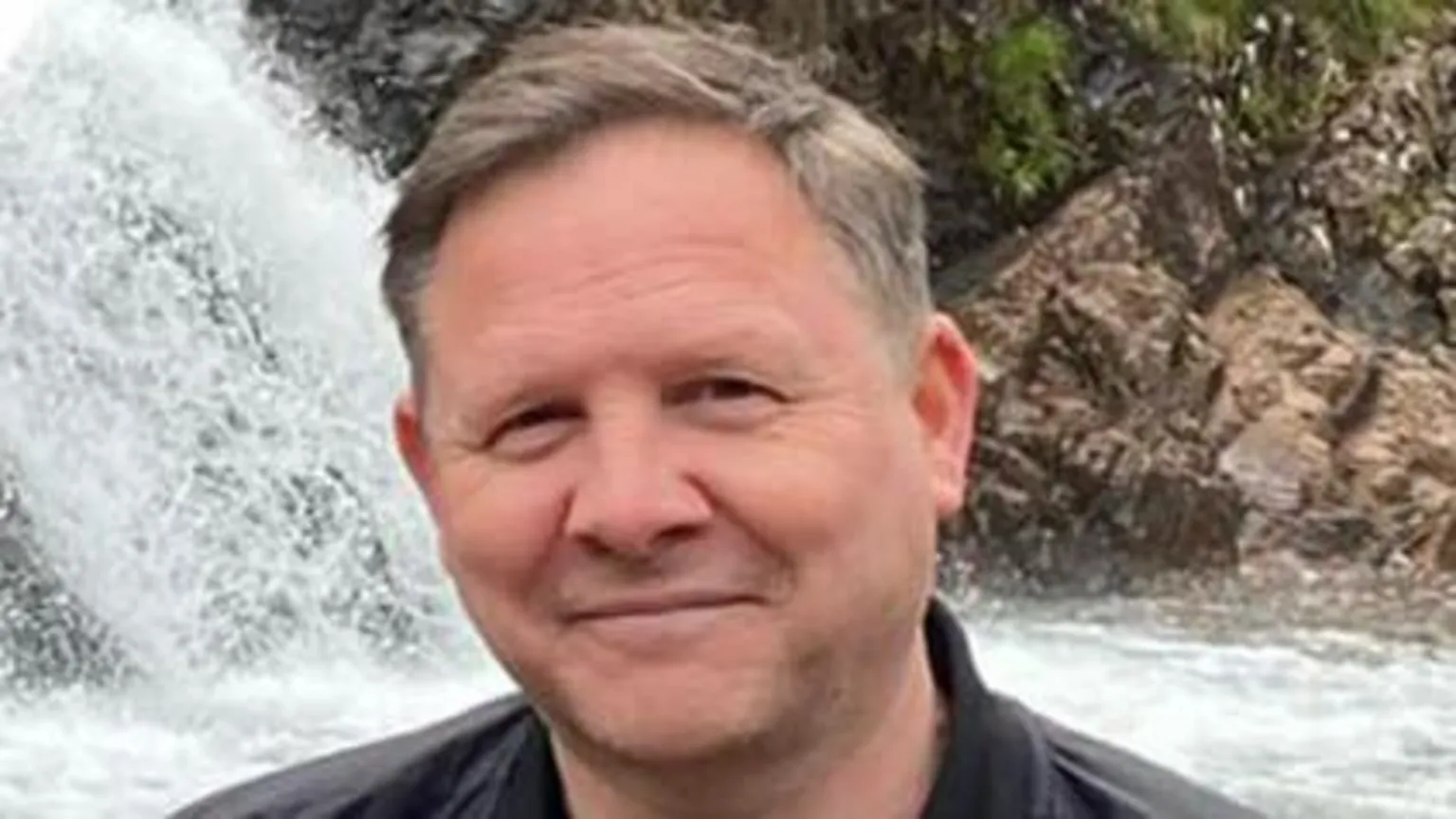 Central Scotland News A man smiling at the camera. He is standing in front of a waterfall. He is wearing a blu et-shirt and a black casual jacket. 