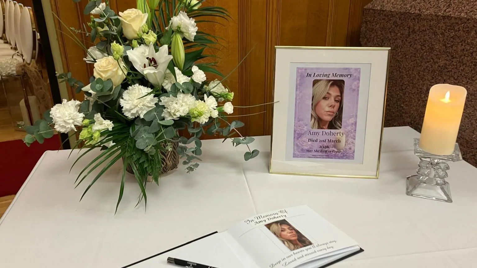 A bunch of flowers and a photograph of a woman are among the items sitting on a table. A book and pen and lit candle can also be seen. All items are atop a white cloth.
