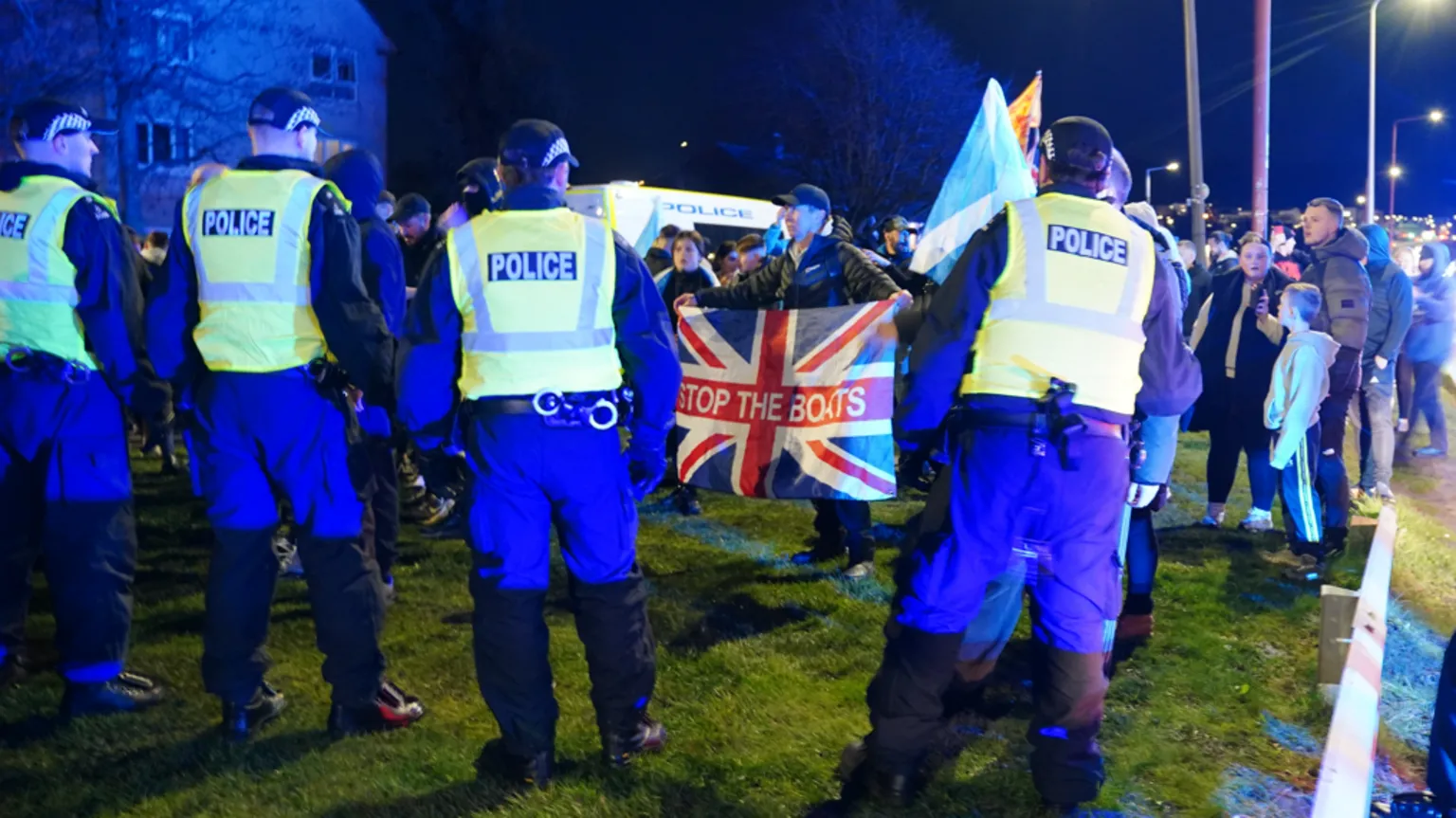  A man holds a union flag with Stop The Boats written on it as he faces four police officers with other protesters behind him