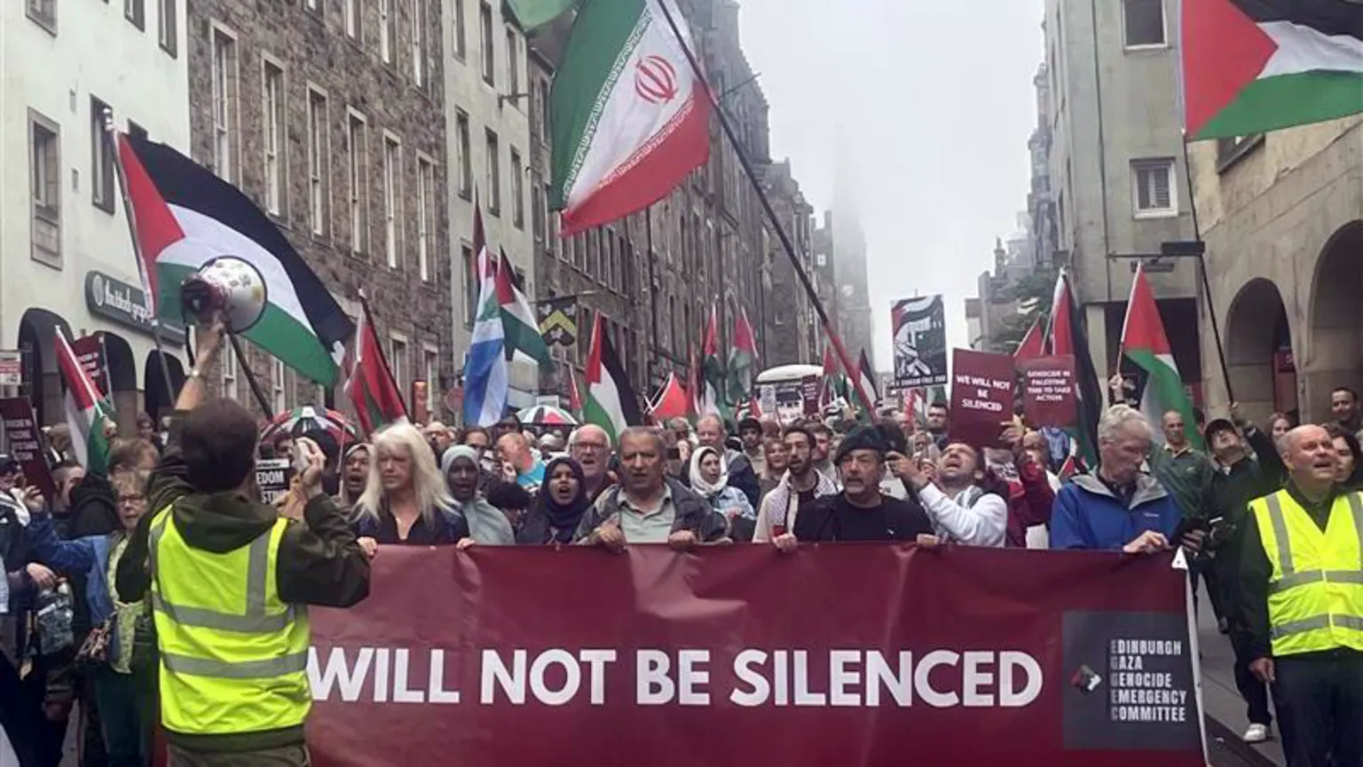 A large protest on the Royal Mile in Edinburgh with several people marching behind a large banner. Some are waving flags of Palestine and Iran. One person has a blue and white Scottish flag.