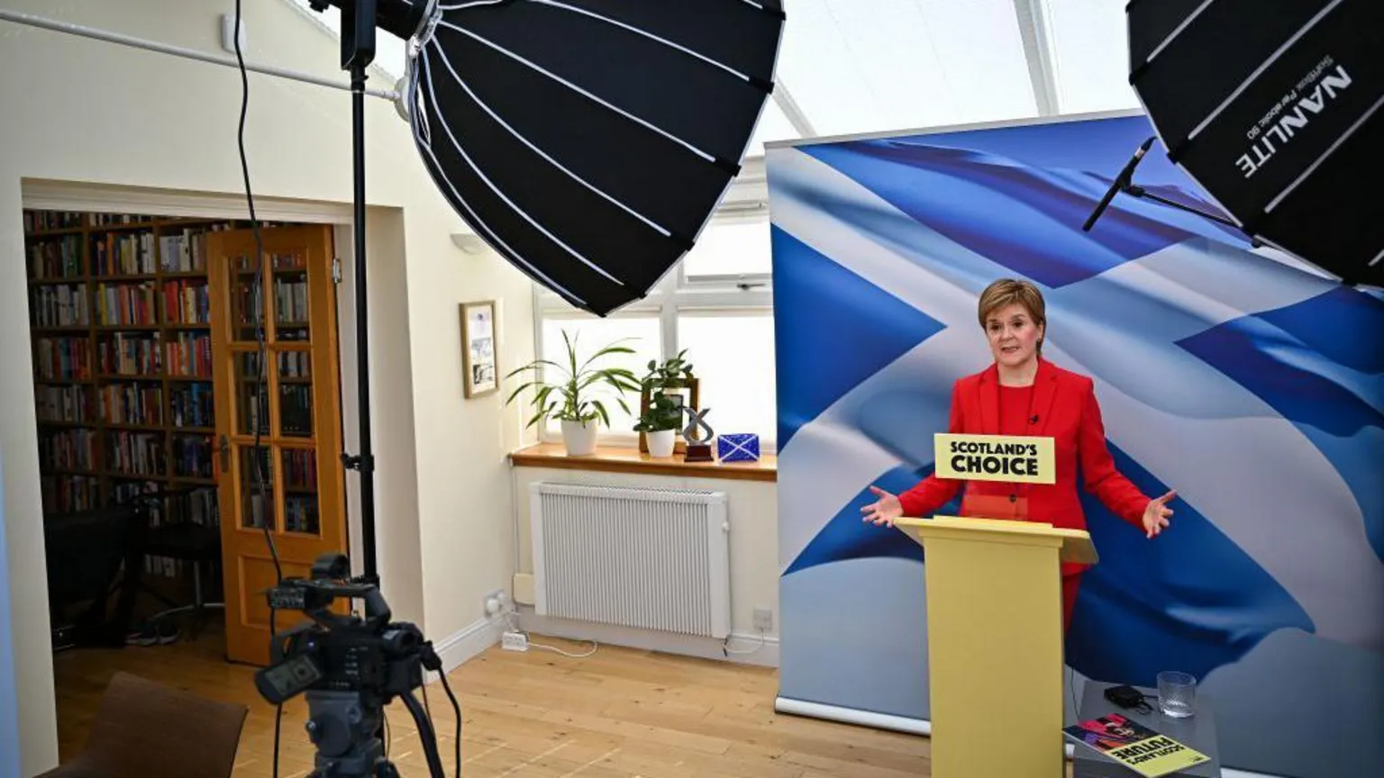  Nicola Sturgeon standing at a podium in her conservatory with a large saltire in the background. She is wearing a red trouser suit.