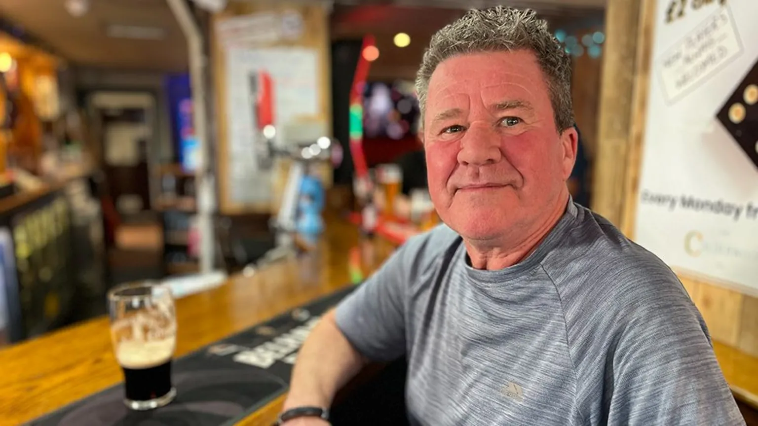 Tam Leddie, who has short grey hair, poses at a bar. He is wearing a grey t-shirt, with a half-full pint on the bar. 