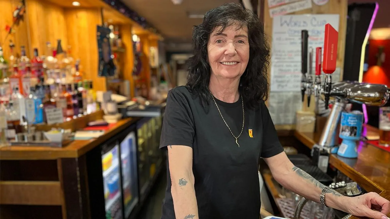 Carol Graham, who has black curly hair, smiles while standing behind a bar. She is wearing a black t-shirt and has tattoos on her forearms. 