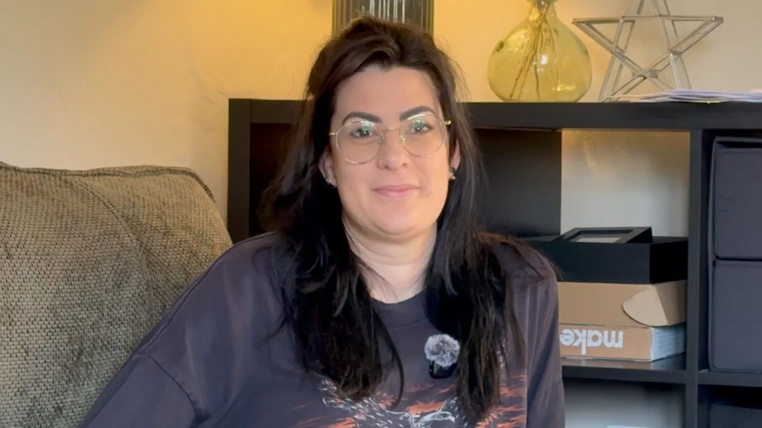 Yolanda Shirin/BBC The woman in the centre has dark brown hair and is sat down. She is wearing wire-framed glasses and is smiling. In the background there is a side table with a vase and a star-shaped ornament on top.