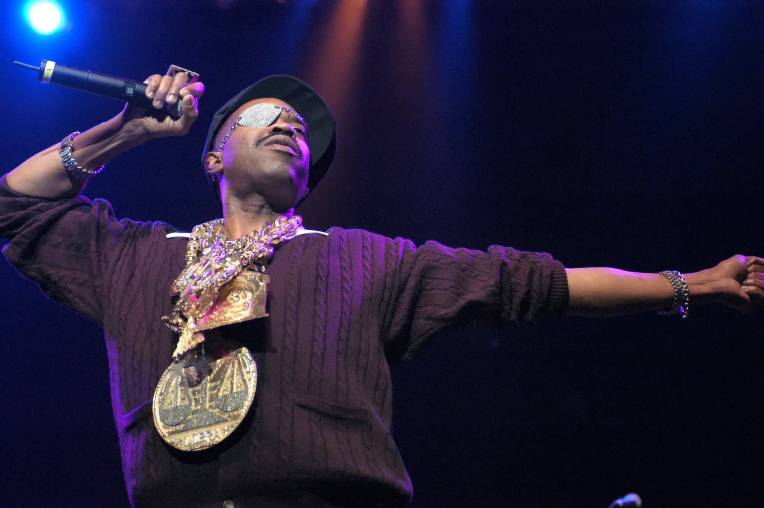  Slick Rick on stage. He is wearing a black baseball cap and a diamond-studded eyepatch, along with a plethora of oversized gold medallions