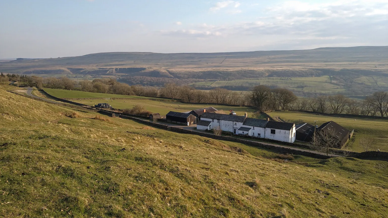 Emma Spry A long white farmhouse in four sections is set at the bottom of a steep hill alongside a single track rural road. Behind it are hills of moorland 