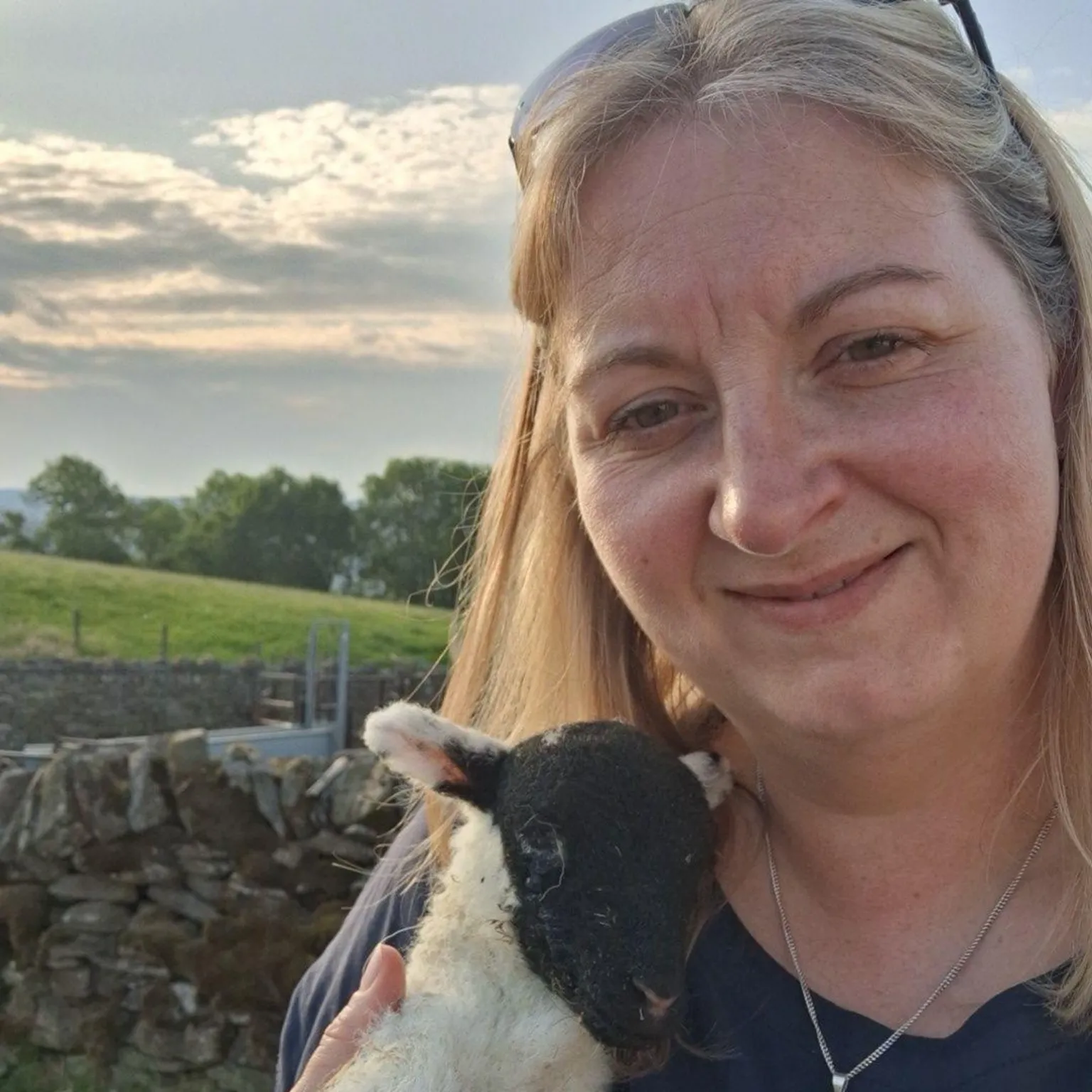 Emma Spry Emma Spry is a woman with long blond hair in her 30s or 40s and is smiling. She is holding a lamb, which has a black face and white body, and there is a field and some trees visible to the left under sunlit clouds.