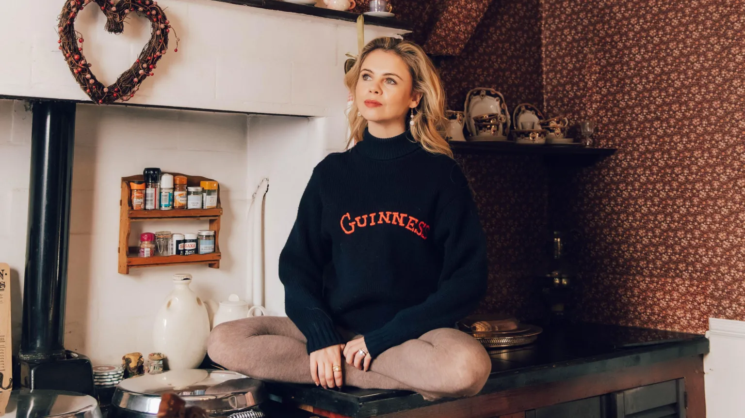 Bryony Coles Saoirse-Monica Jackson has long blonde curly hair. She is sitting on a kitchen bench and wearing a black jumper with the word Guinness written in red block letters.