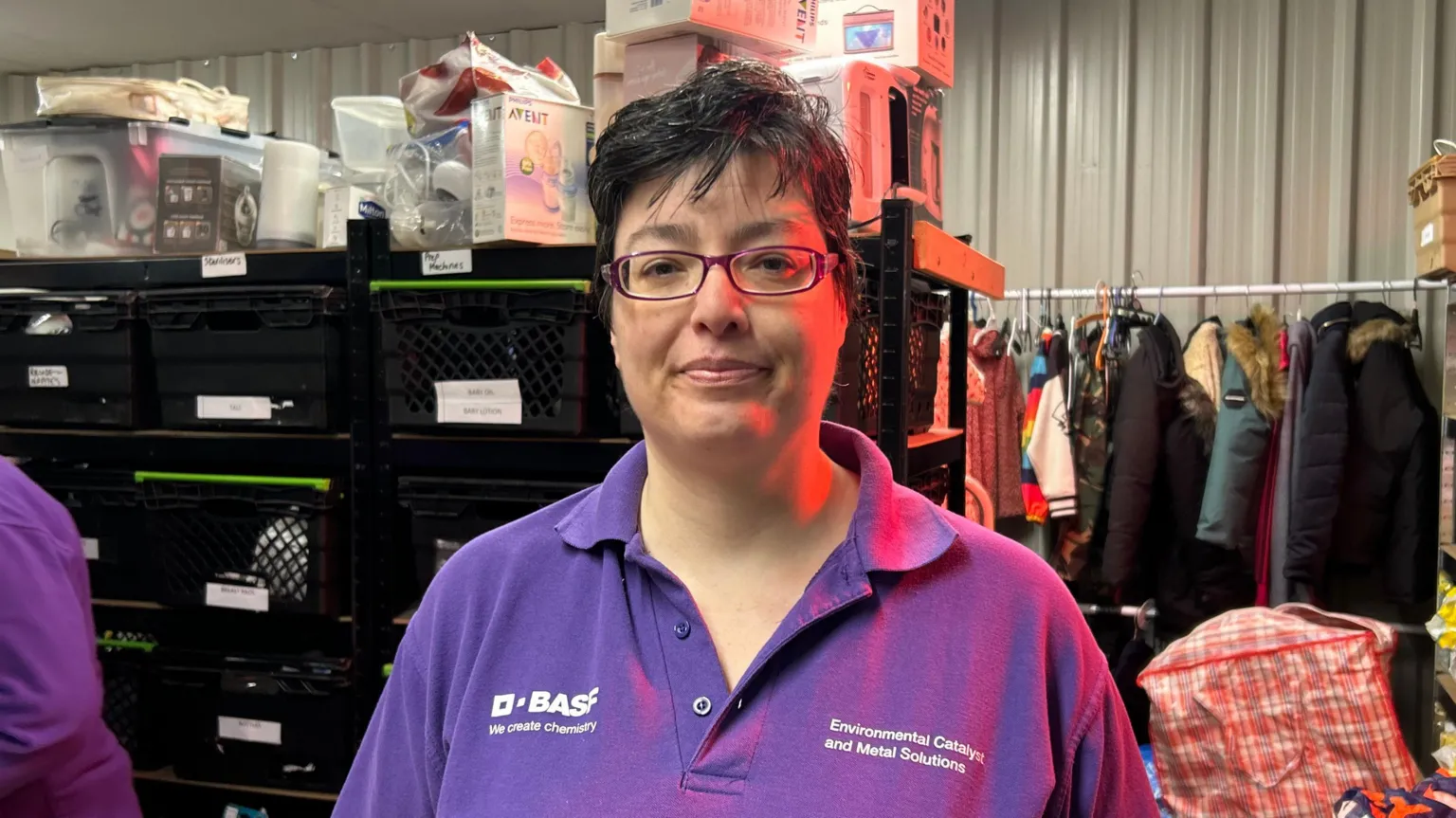 A photo of a woman stood in front of a storage area. She is wearing a purple top and has dark hair and is wearing glasses