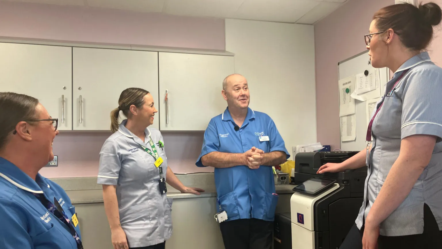 Fraser Morton pictured chatting with three female colleagues in a staff or storage room within a hospital setting. He is wearing a blue midwife's tunic with identification badges. He is leaning against a cupboard and his hands are clasped in front of his chest. The women are all dressed in healthworker's tunics. The group are smiling as they chat.