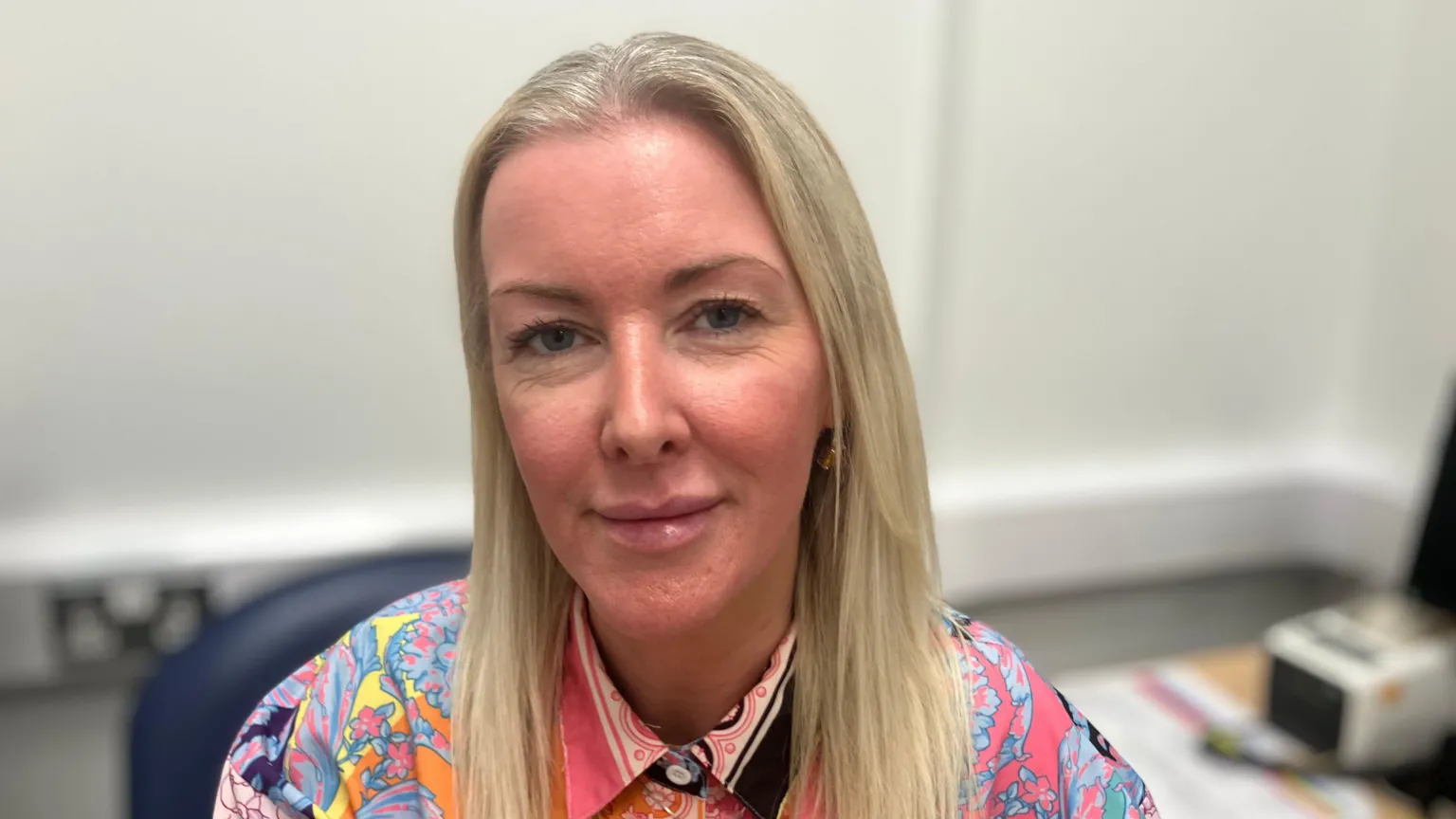 Laura Rooney, a woman with long, straight blonde hair, smiles at the camera while sitting in an office. She is wearing a bright, multicoloured patterned blouse buttoned up to the neck. There is a computer in the background.