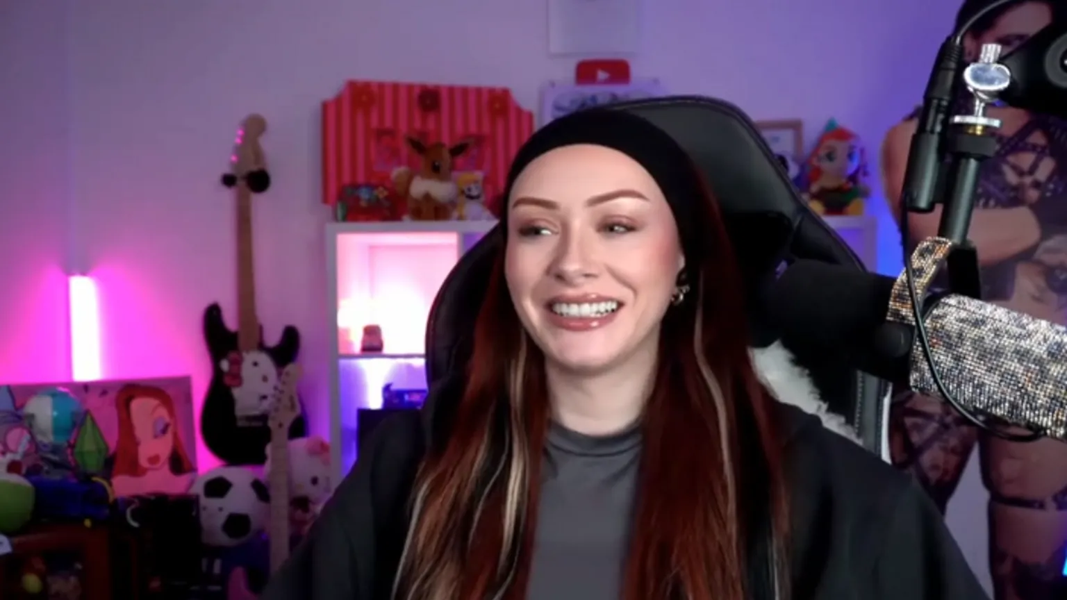 Elz is wearing a black headband and a black open hoodie with a grey t-shirt underneath. She is sat at a gaming char with a silver sparkly mic to her left.