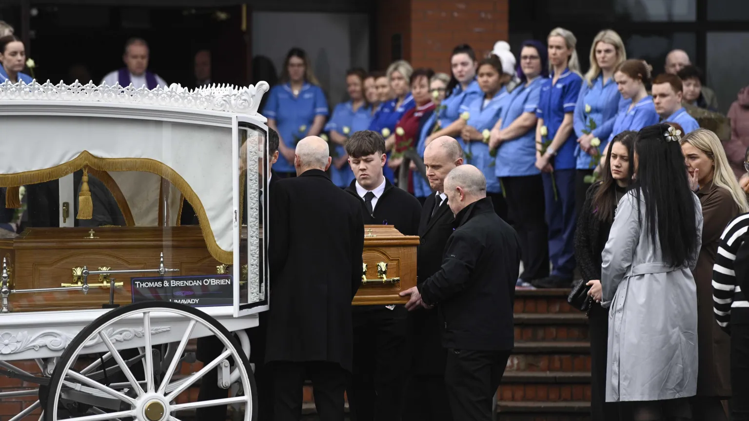 A white carriage carrying a coffin sits at the bottom of a set of red brick steps. Gathered around it are mourners, some dressed in black and others in the sky and darker blue of the NHS.