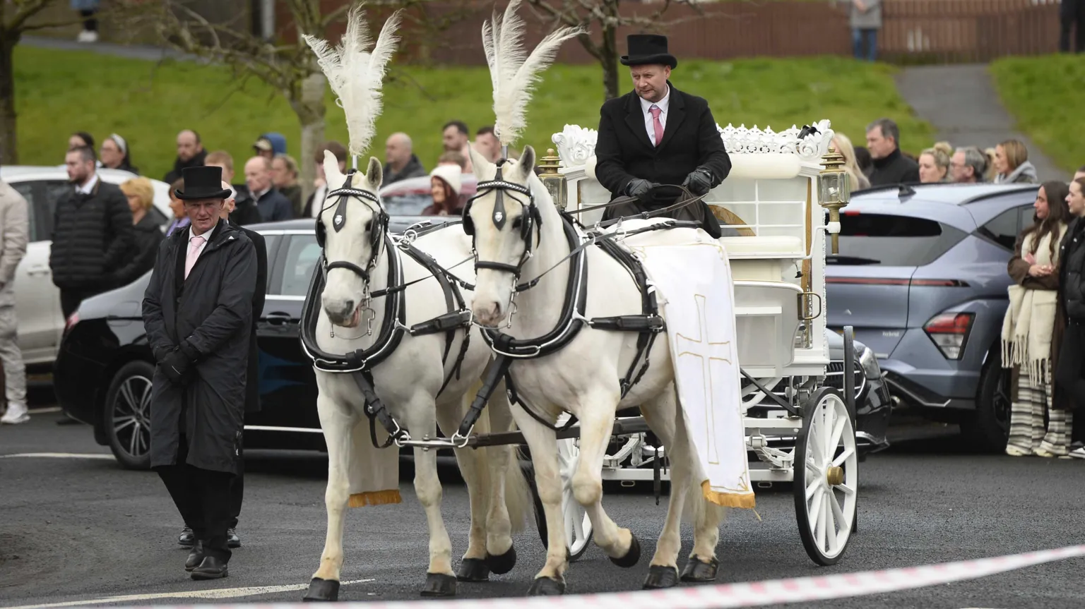 Two white horses wearing funeral gear pull a white carriage carrying Amy's coffin. Behind the horses a man holds the reigns, and beside them walks another funeral director, dressed in black, hands clasped solemnly.
