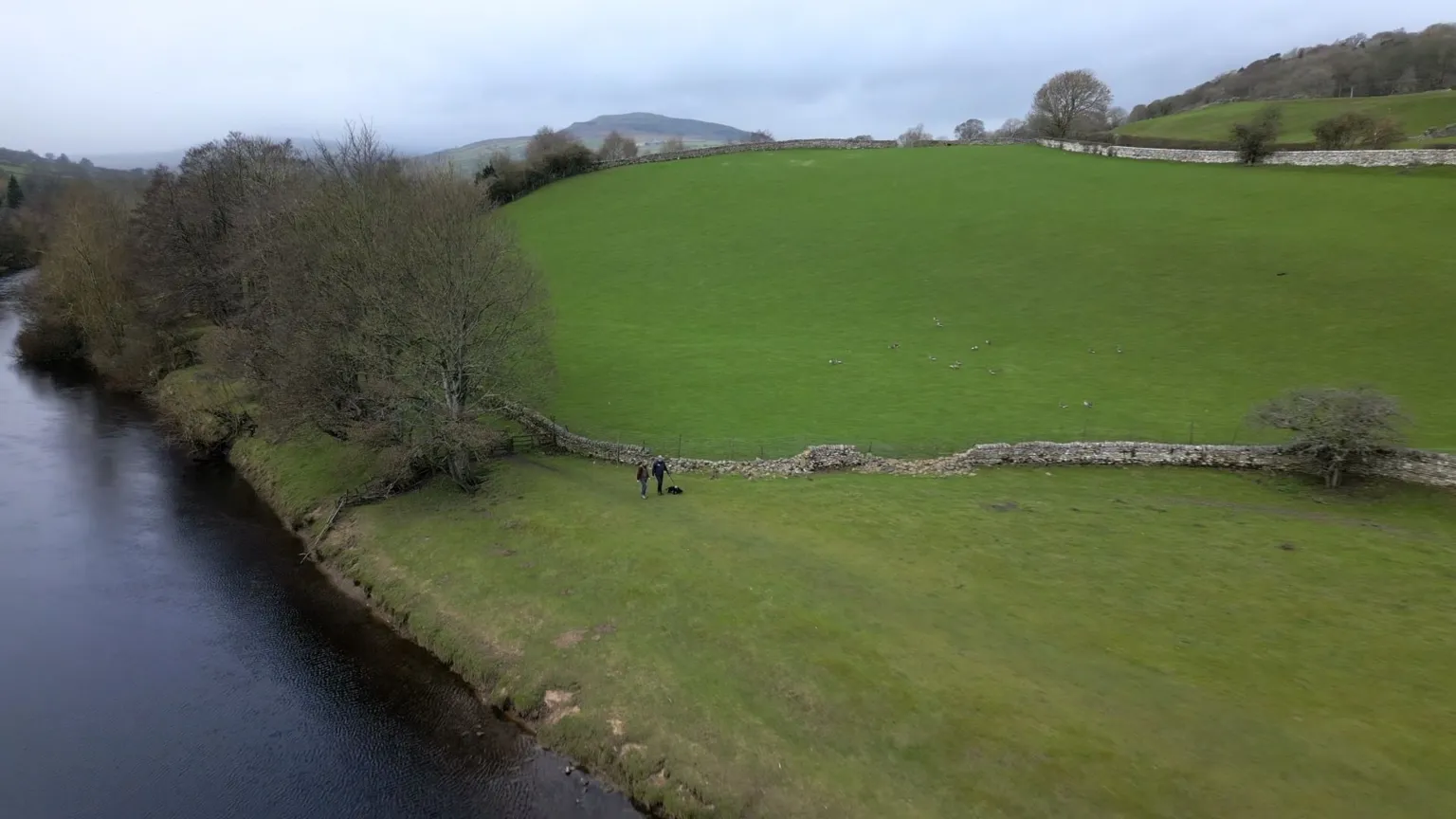 Swaledale in the Yorkshire Dales National Park . Two people, Danny and Mark , and a dog stand on a grassy riverbank beside a wide, calm river. A line of trees borders the water, and an old stone wall curves across the field behind them. The landscape rises into green hills with scattered trees and additional stone walls, leading towards distant higher ground under an overcast sky.