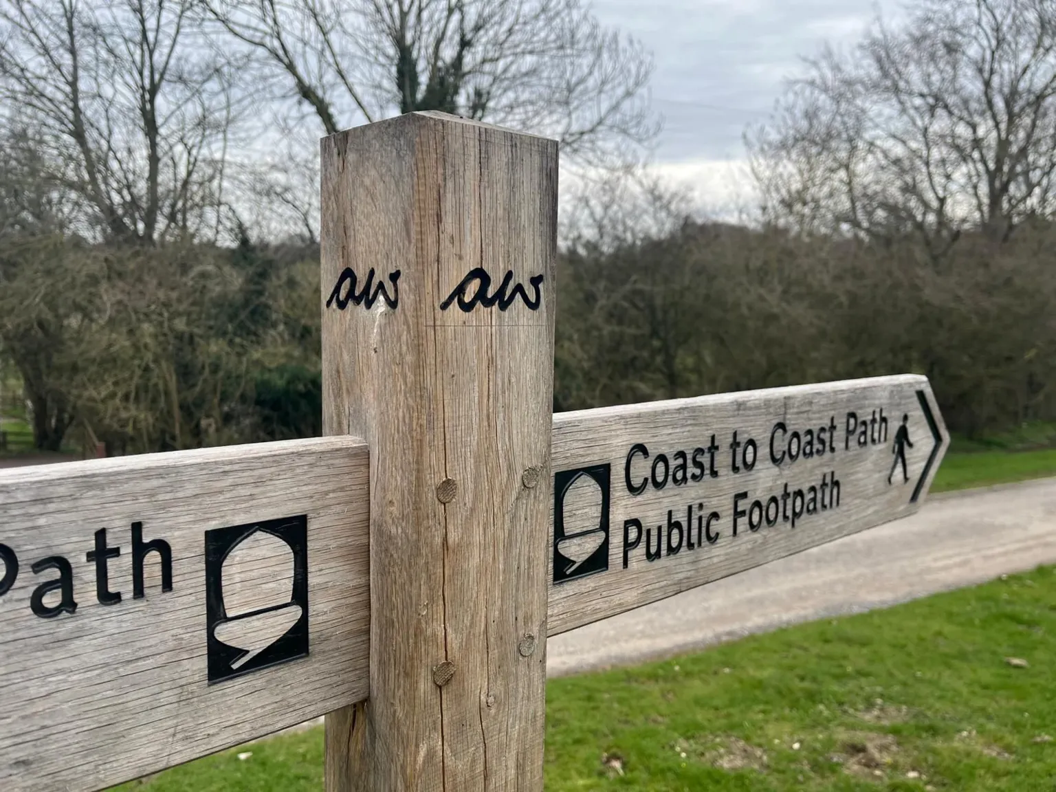 A wooden signpost stands at a junction of paths, with two horizontal arms pointing in different directions. The right‑hand arm reads “Coast to Coast Path, Public Footpath” with a walking‑figure symbol. The left‑hand arm shows part of the same acorn‑shaped National Trail marker. Trees and grassland fill the background, and a gravel path runs behind the sign. In the centre of the sign post are the initials aw carved into it.