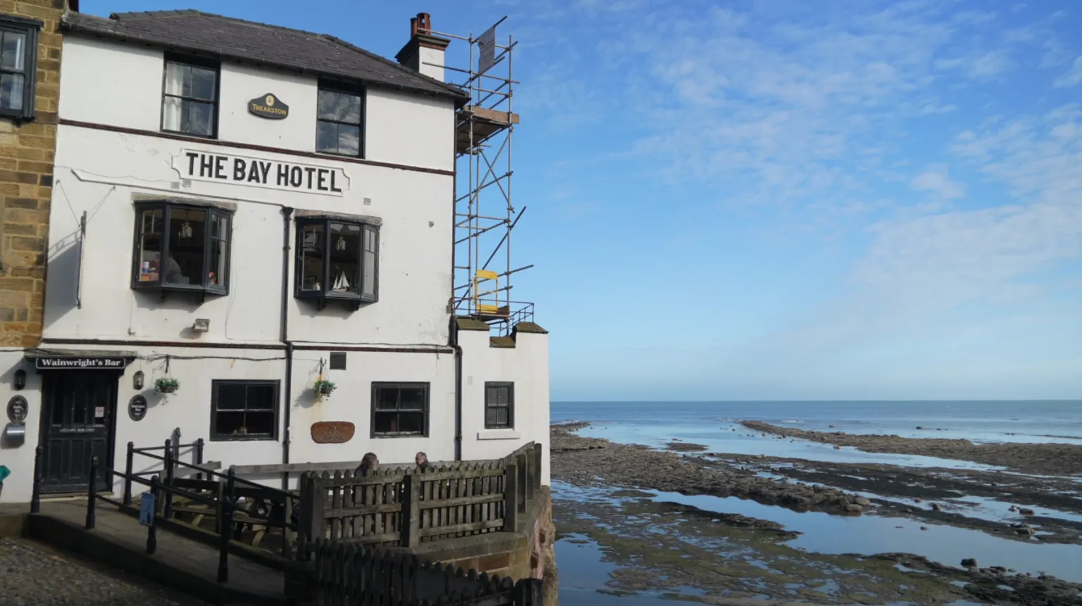 A white seaside building labelled “The Bay Hotel” stands on a cobbled slope beside the shoreline. The building has multiple windows, a doorway to a bar, and scaffolding on its right side. A wooden fence runs along the front. To the right, the rocky beach is exposed at low tide, with shallow pools of water leading out to the calm sea beneath a blue, lightly clouded sky.
