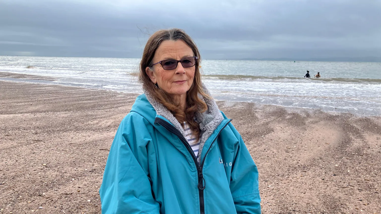 A woman in a blue waterproof coat stands on the beach in Exmouth, Devon. There are two people in the sea behind her. You can see a stretch of sand. The sky is cloudy. 