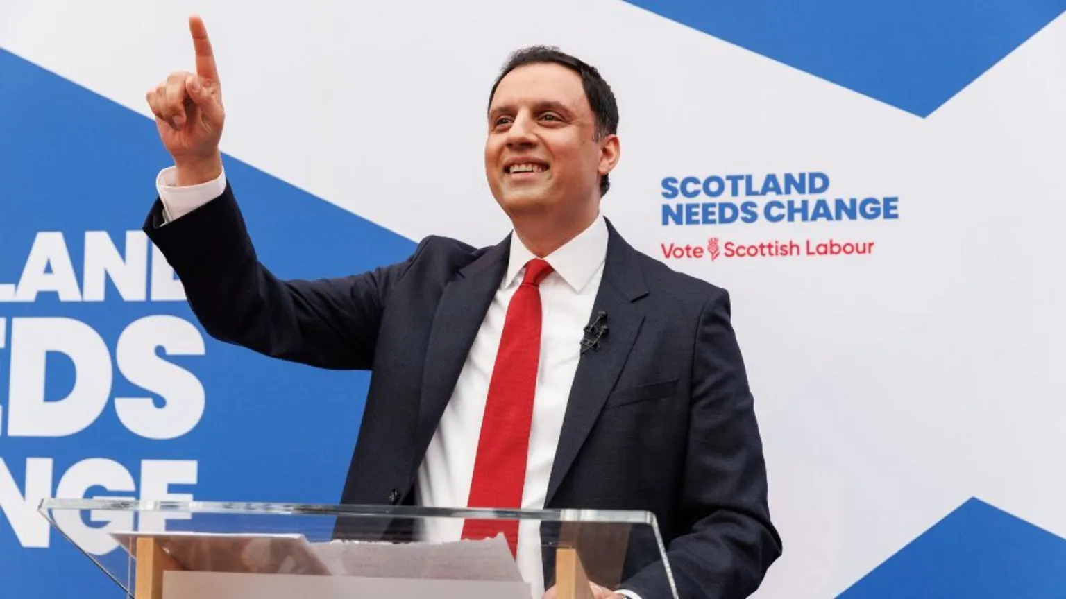  Anas Sarwar, who has short black hair, in front of a saltire background. He is wearing a business suit and a red tie, pointing in the air and smiling. 