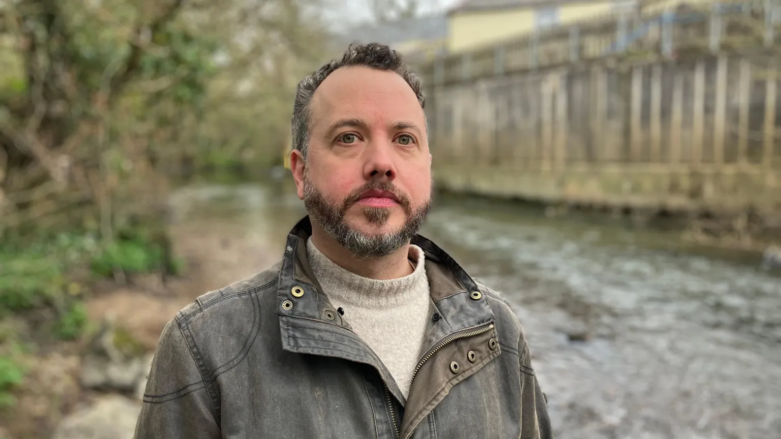 A man in a green waterproof jacket stands by the River Lew at Hatherleigh 