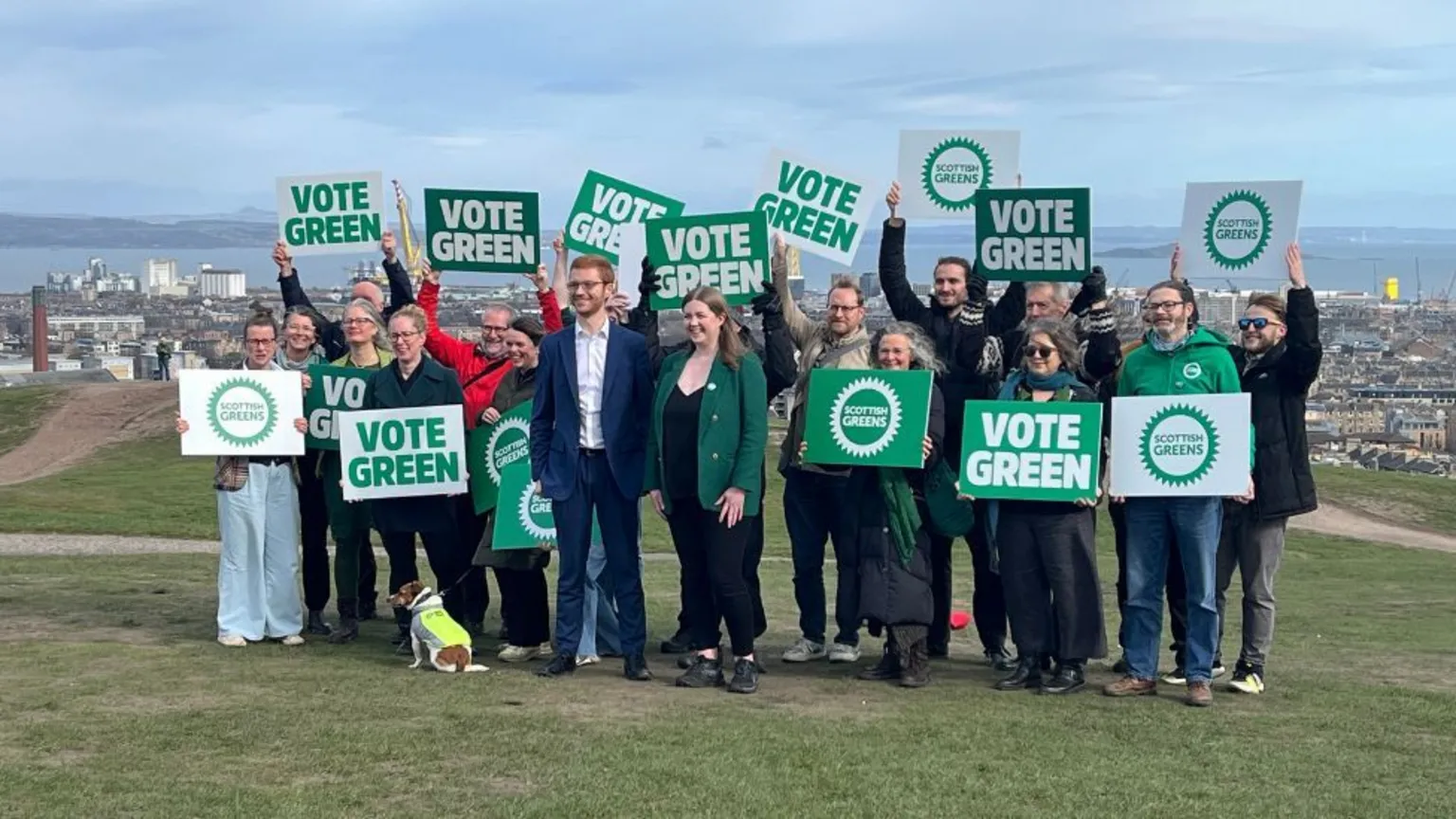 A group of smiling people holding Green Party placards at the top of a grassy hill overlooking a city 
