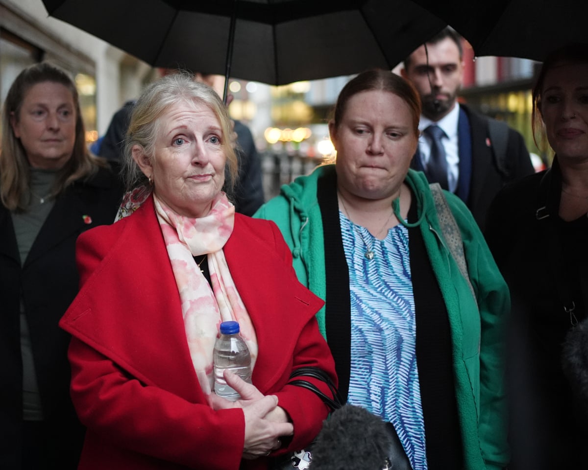 A woman in a red coat stands next to a woman in a green coat, talking to the media. They have pained, sad expressions.
