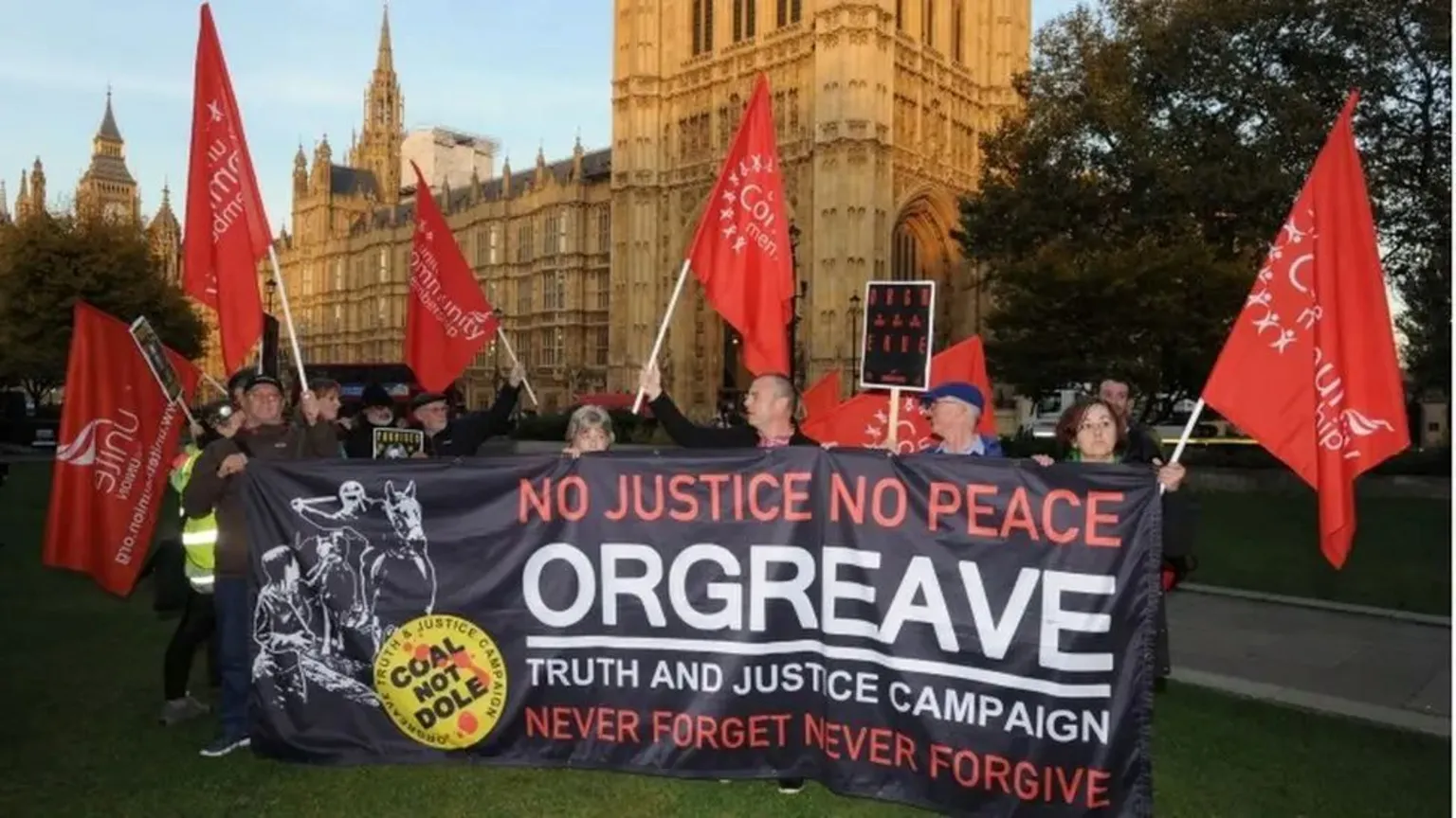 A crowd of people are standing outside the House of Commons with a large black, orange and white banner saying