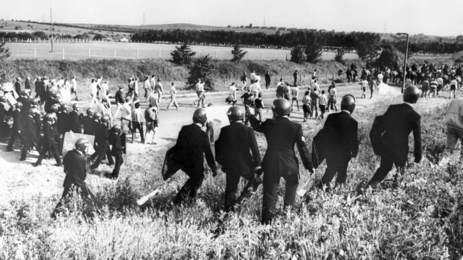 A black and white photo shows a line of police officers with shields confronting miners in normal clothes in a field.