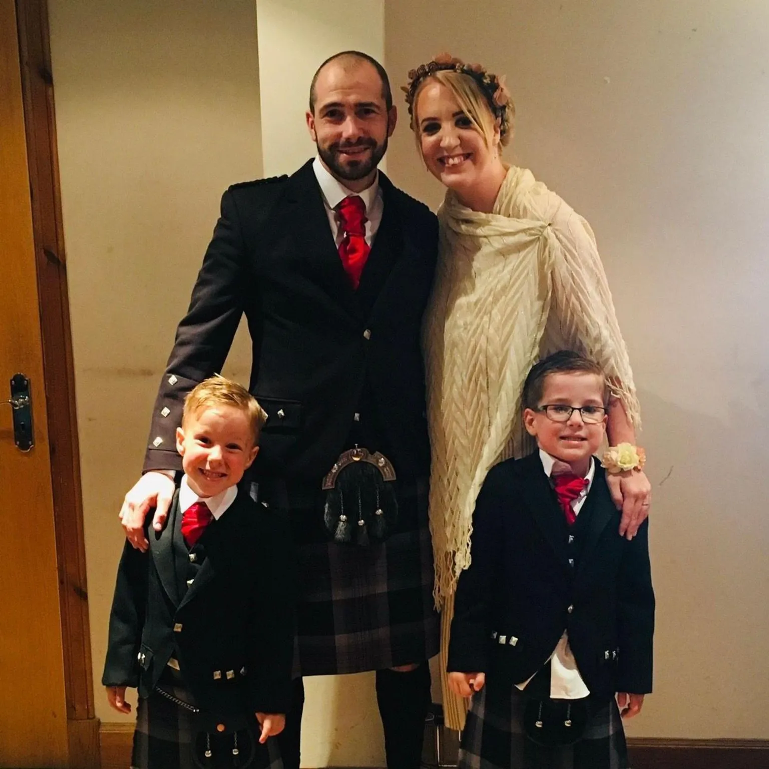 Shelley Martin Barry is wearing a kilt as are his sons. His wife is wearing a white shawl and has her hair up with a wreath of flowers in it.