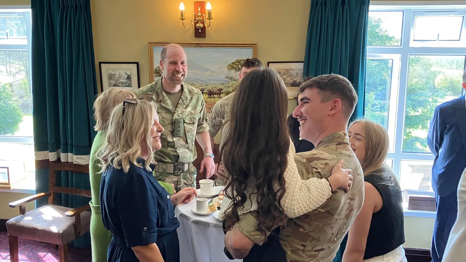 Will Glennon People stood around a white table smiling with cups of tea in front of them. Prince William is among them and his smiling at a man who is holding his young daughter, who has long dark brown hair