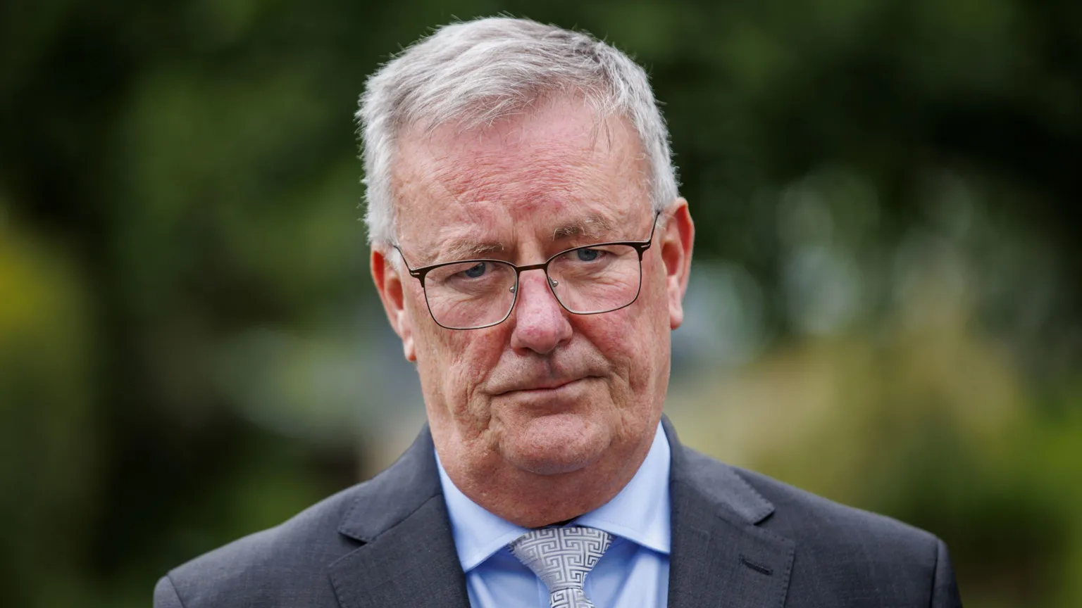 Mike Nesbitt is wearing a blue shirt, white tie and grey blazer. He has short grey hair and glasses on his face. The background is blurry.