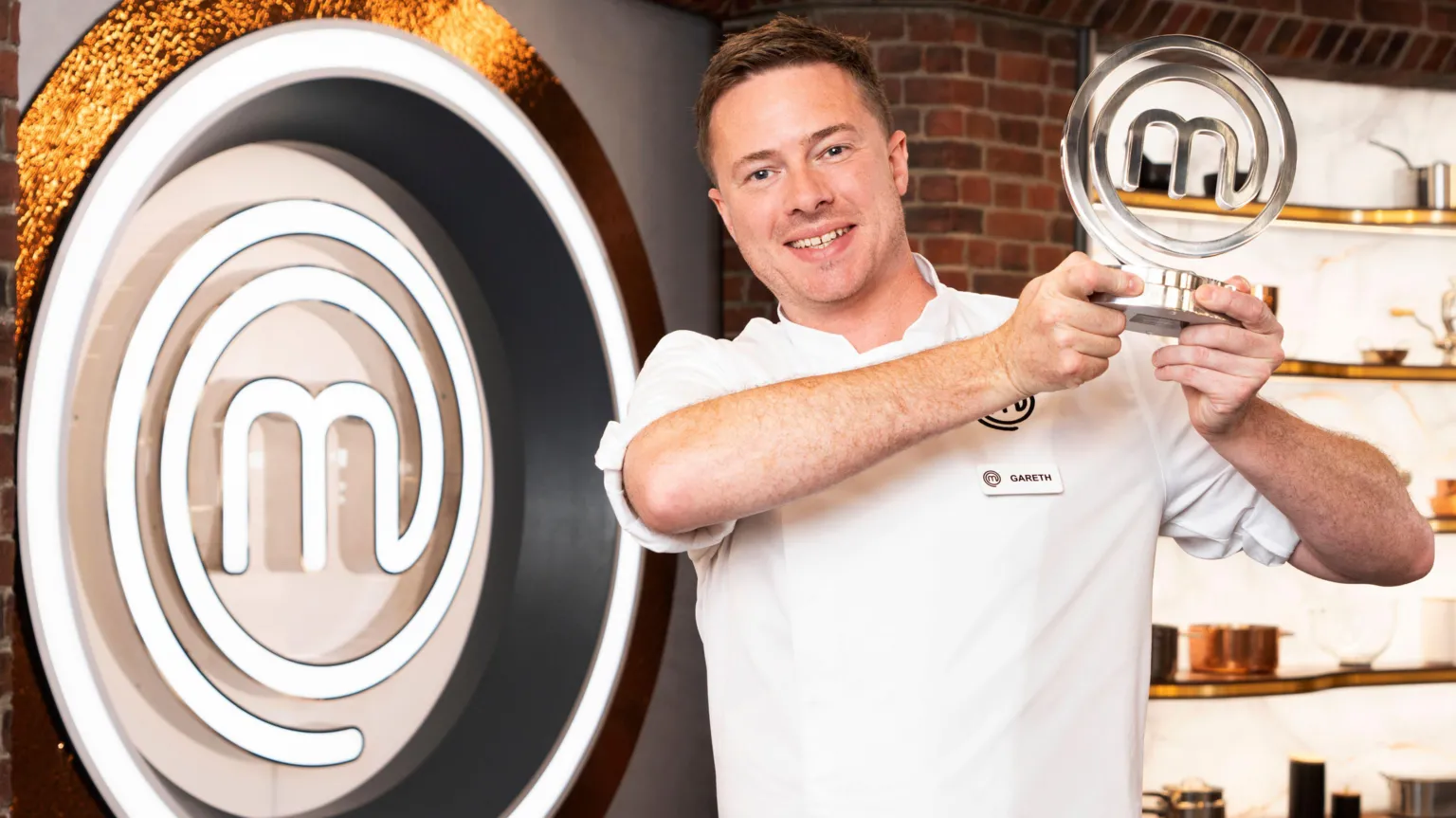 BBC/Shine TV Gareth Baty holds up the MasterChef trophy on the set of the TV show. The logo of the show takes up the whole wall next to him, while behind him are some pans and utensils on shelbes. He has short brown hair and blue eyes. He is wearing a white chef's coat and smiling at the camera.