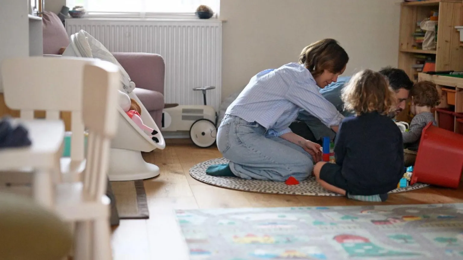 The family sits on the floor gathered around a small play area with building blocks. The room contains children’s furniture, shelves with storage boxes and toys, a rug with colourful patterns and a ride‑on toy near the window.