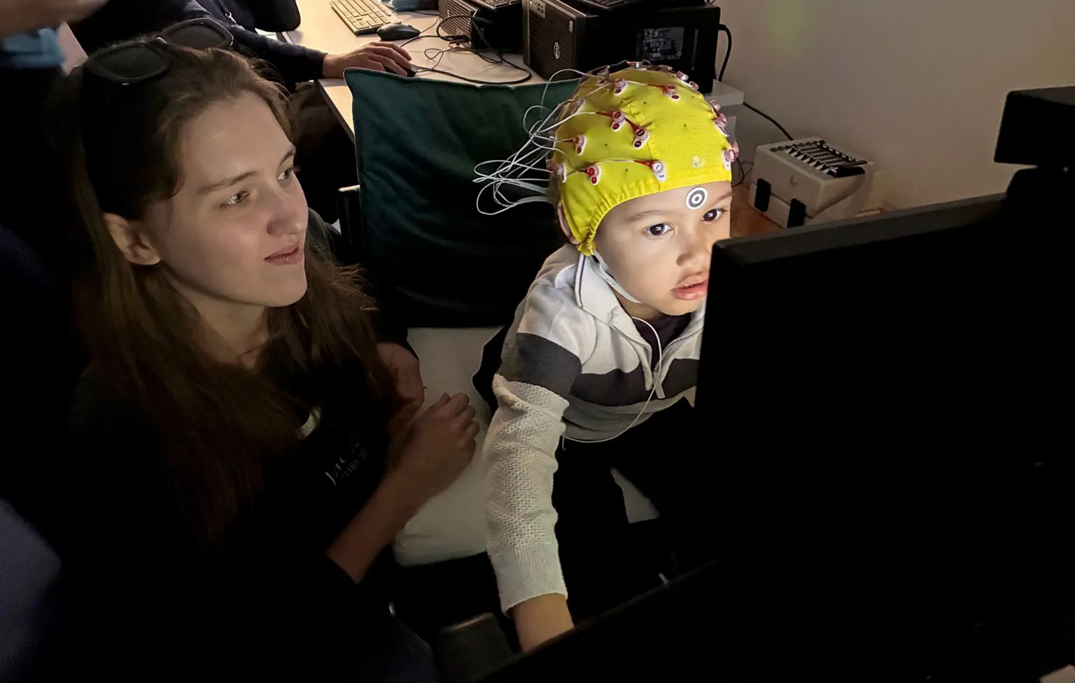 A young boy wearing a yellow cap fitted with multiple wires sits at a computer workstation and watches a video while engaged in a research activity. Behind, computer equipment and monitors fill the dimly lit room and one of the researchers looks on in frame.