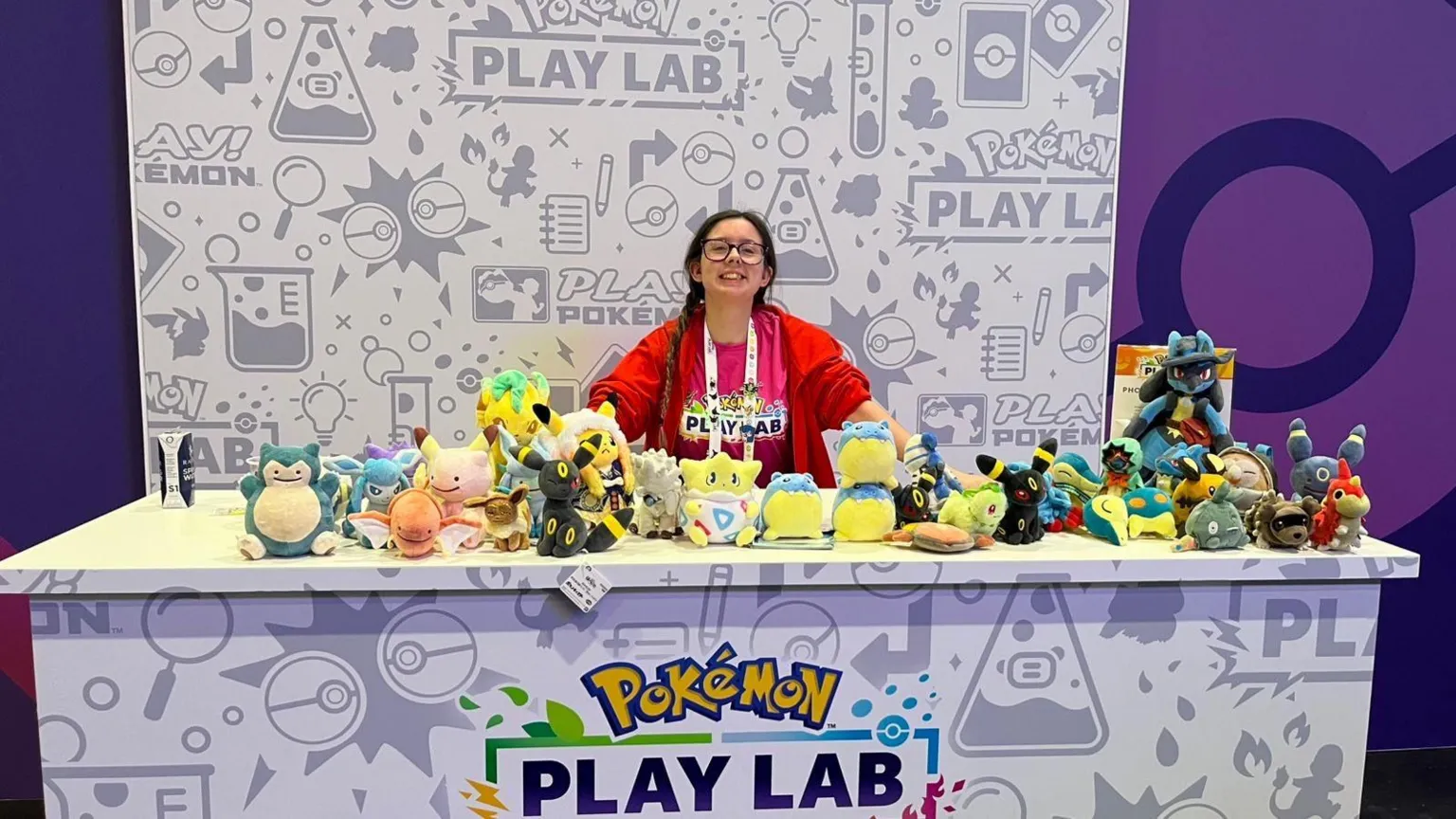 Morgan Elise A brunette girl in a red jacket is stood smiling behind a branded panel full of colourful Pokémon plushies.
