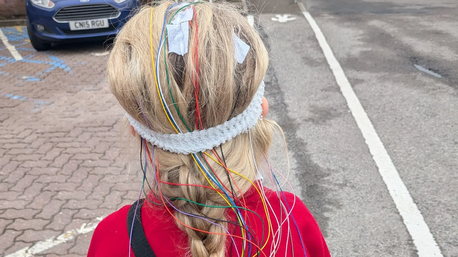 Emma Alderman The behind of Noa-Rose's head which has many multi-coloured wires coming out of. She is stood in a car park.