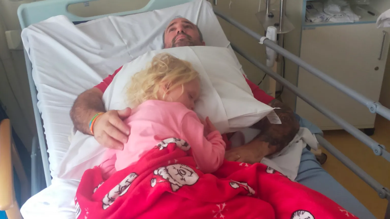 Emma Alderman Noa-Rose sleeping on a pillow on top of Scott in a hospital bed. Noa-Rose wears pink pyjamas and has a red blanket on op of her. Scott has his arm around her and stares up to the ceiling with a worried look.