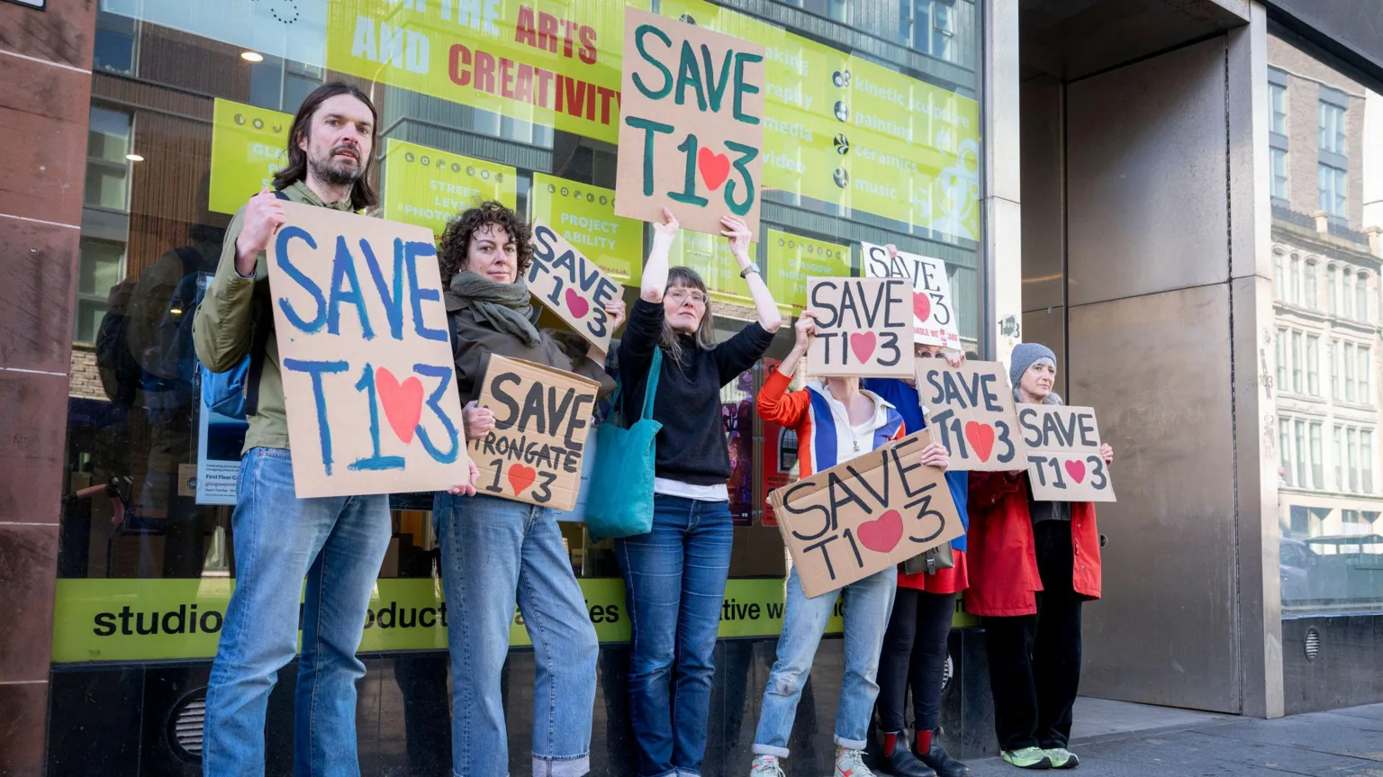 Kirsty Anderson Six people holding placards reading Save T103, except the 0 is an heart. They are all leaning against the wall of a large building.