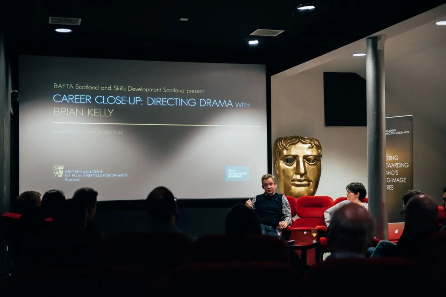  A group of people in a small cinema watching a talk or in conversation event. The big screen shows CAREER CLOSE-UP: DIRECTING DRAMA as the headline.