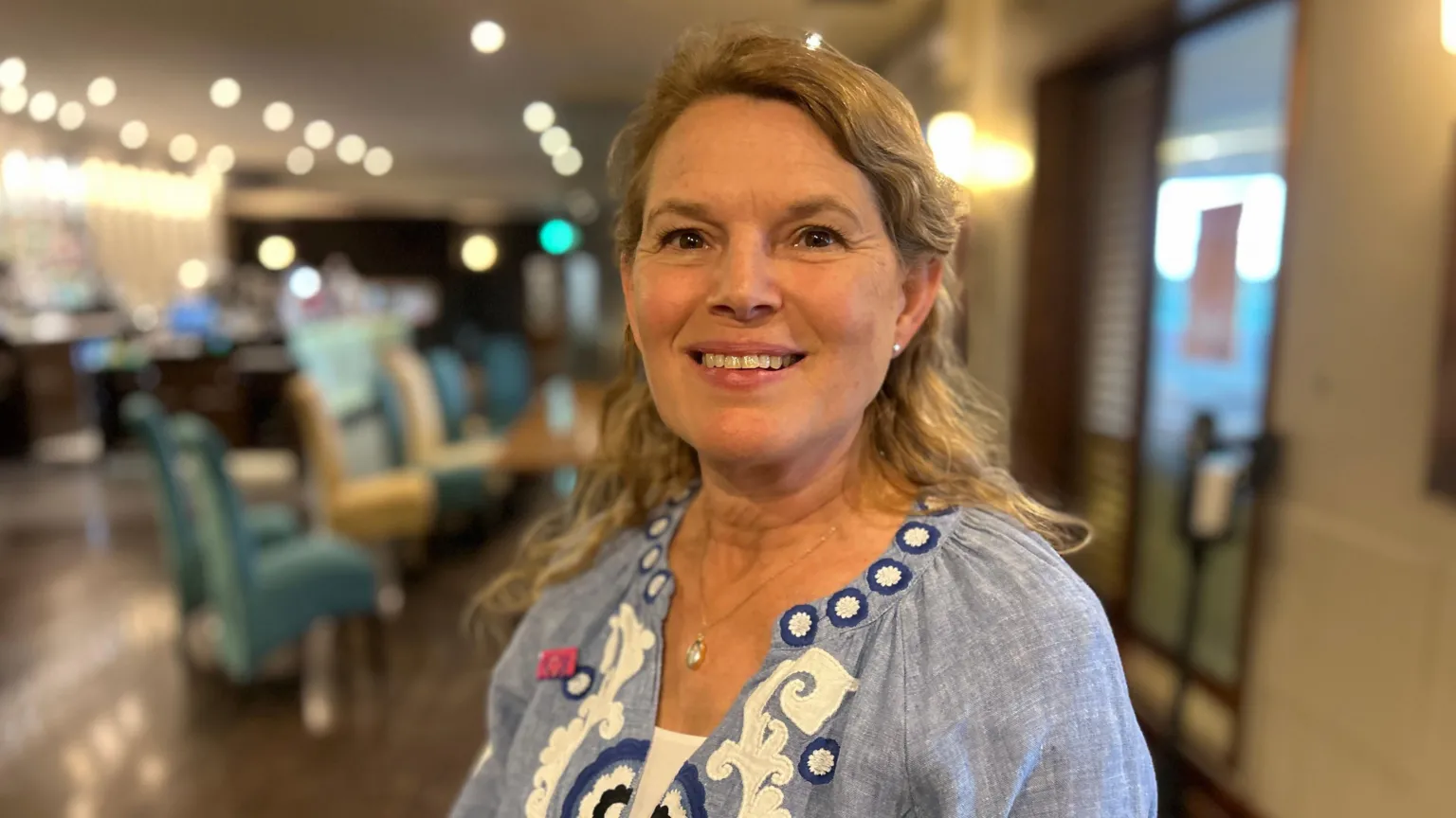 A woman with long, blonde, wavy hair in a blue patterned blouse stand in front of what looks like a blurred hotel bar area 
