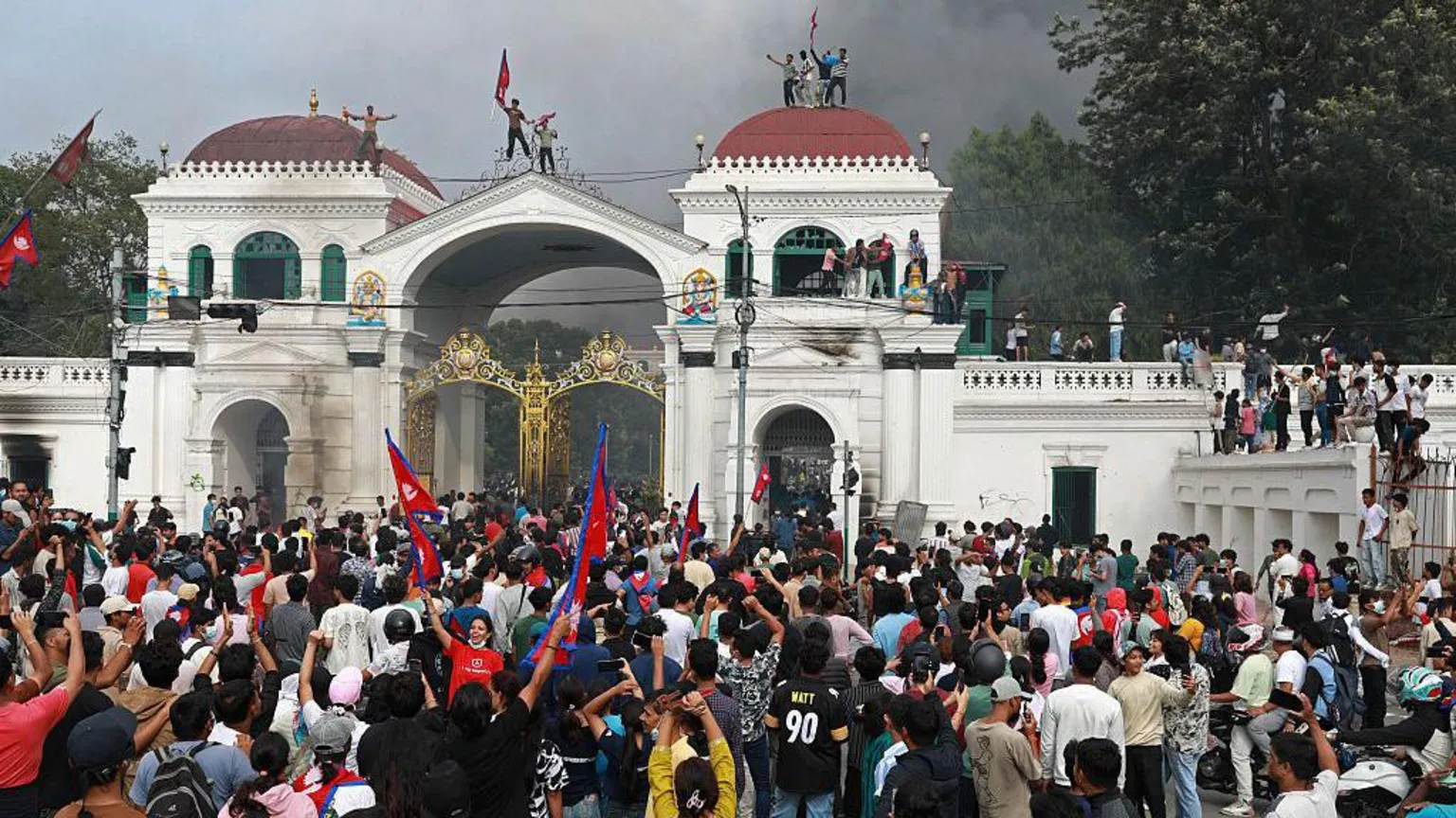 Hundreds of protesters gather at the Singha Durbar, the main administrative building for the Nepal government. Some are seen waving the national flag and some are standing on the roofs of the building. A cloud of smoke is billowing in the background